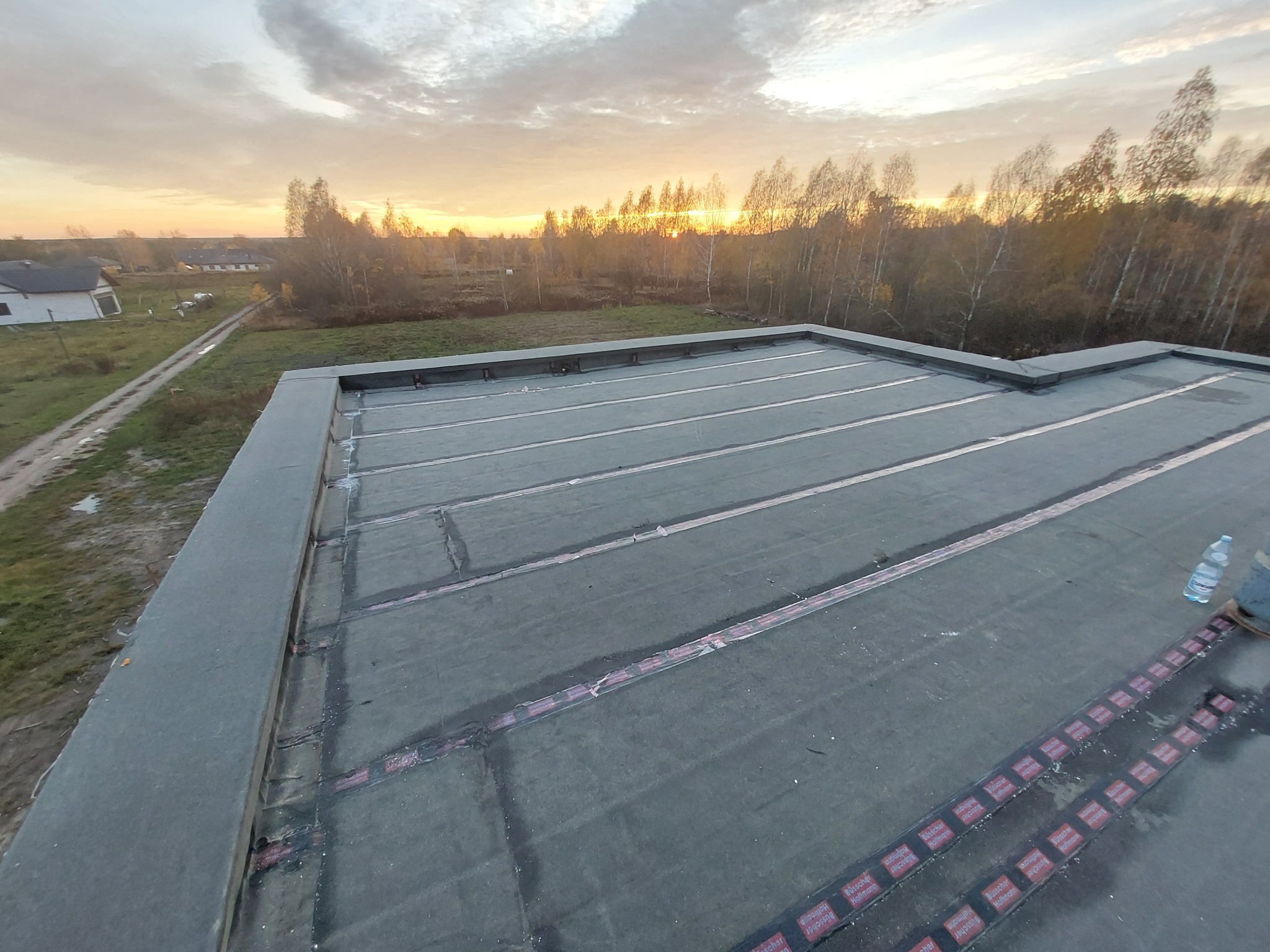 Płaski dach pokryty papą termozgrzewalną z widocznymi pasami łączeń i butelką wody w rogu, na tle zachodzącego słońca i jesiennego krajobrazu.