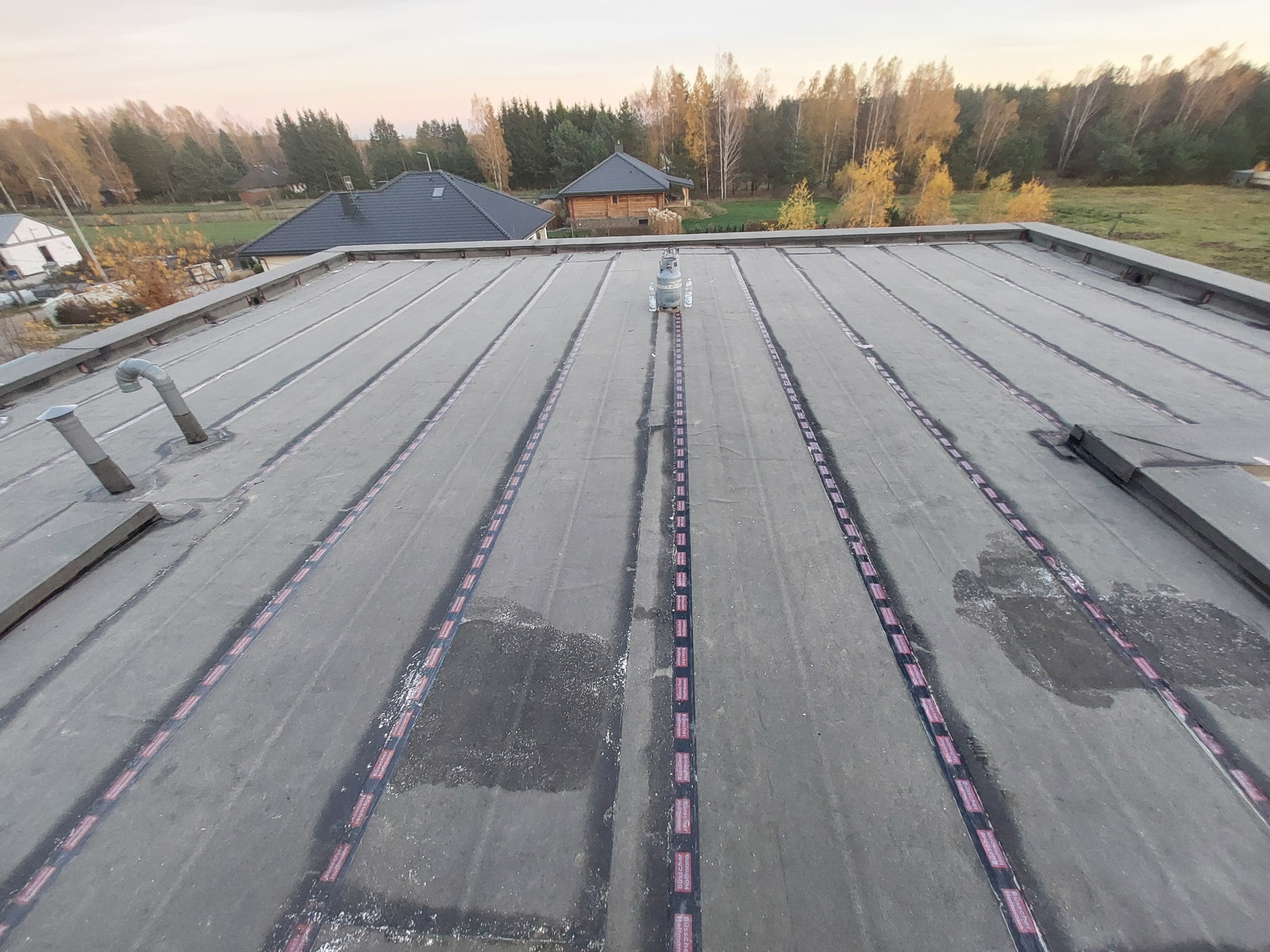 Szary, płaski dach pokryty papą termozgrzewalną z widocznymi zgrzewami. W tle budynki i drzewa. Na dachu butle z gazem.