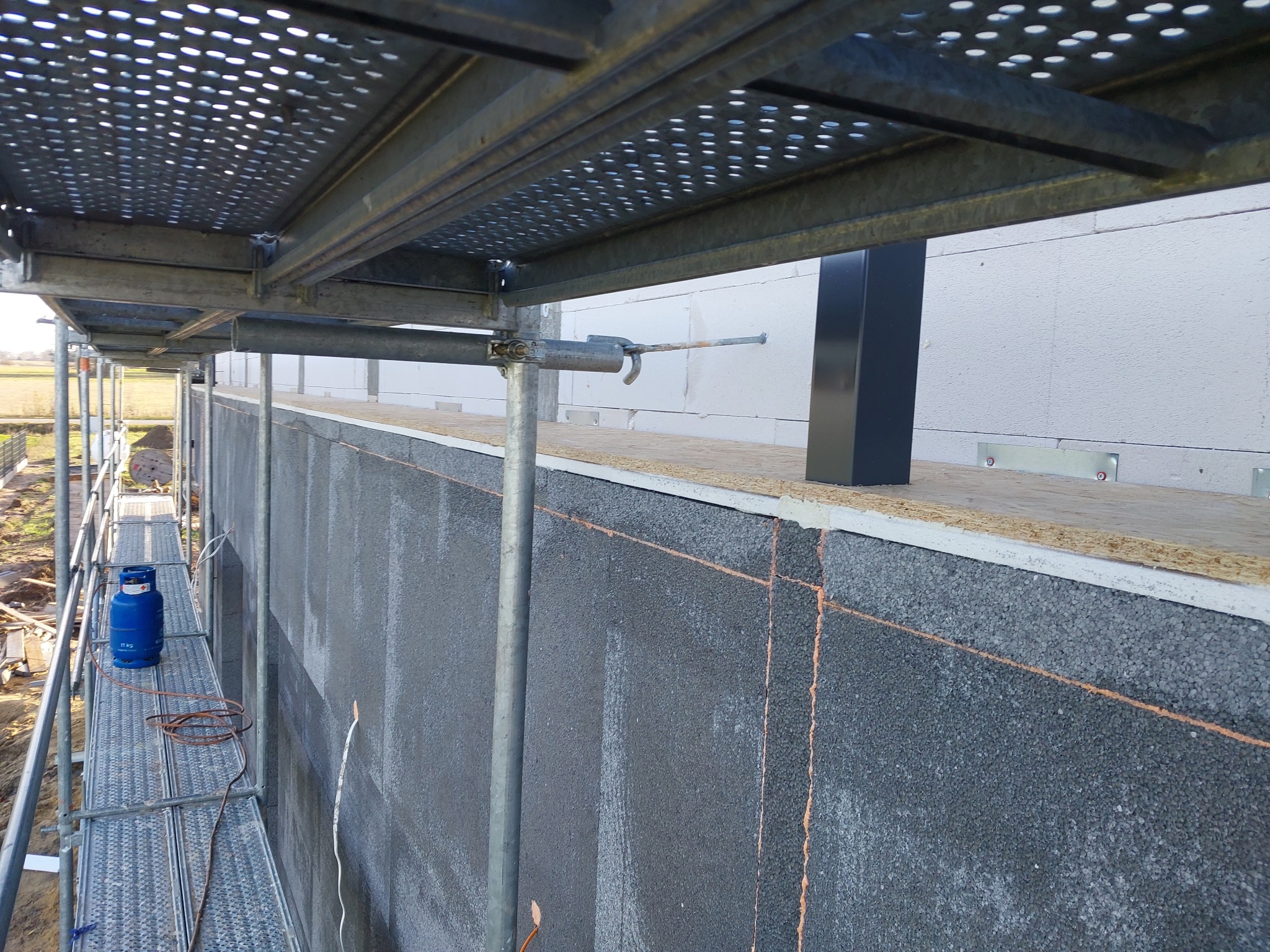 Ocieplanie ściany zewnętrznej budynku szarym styropianem grafitowym, widoczne rusztowanie i butla gazowa. Prace izolacyjne w toku, detale montażu.