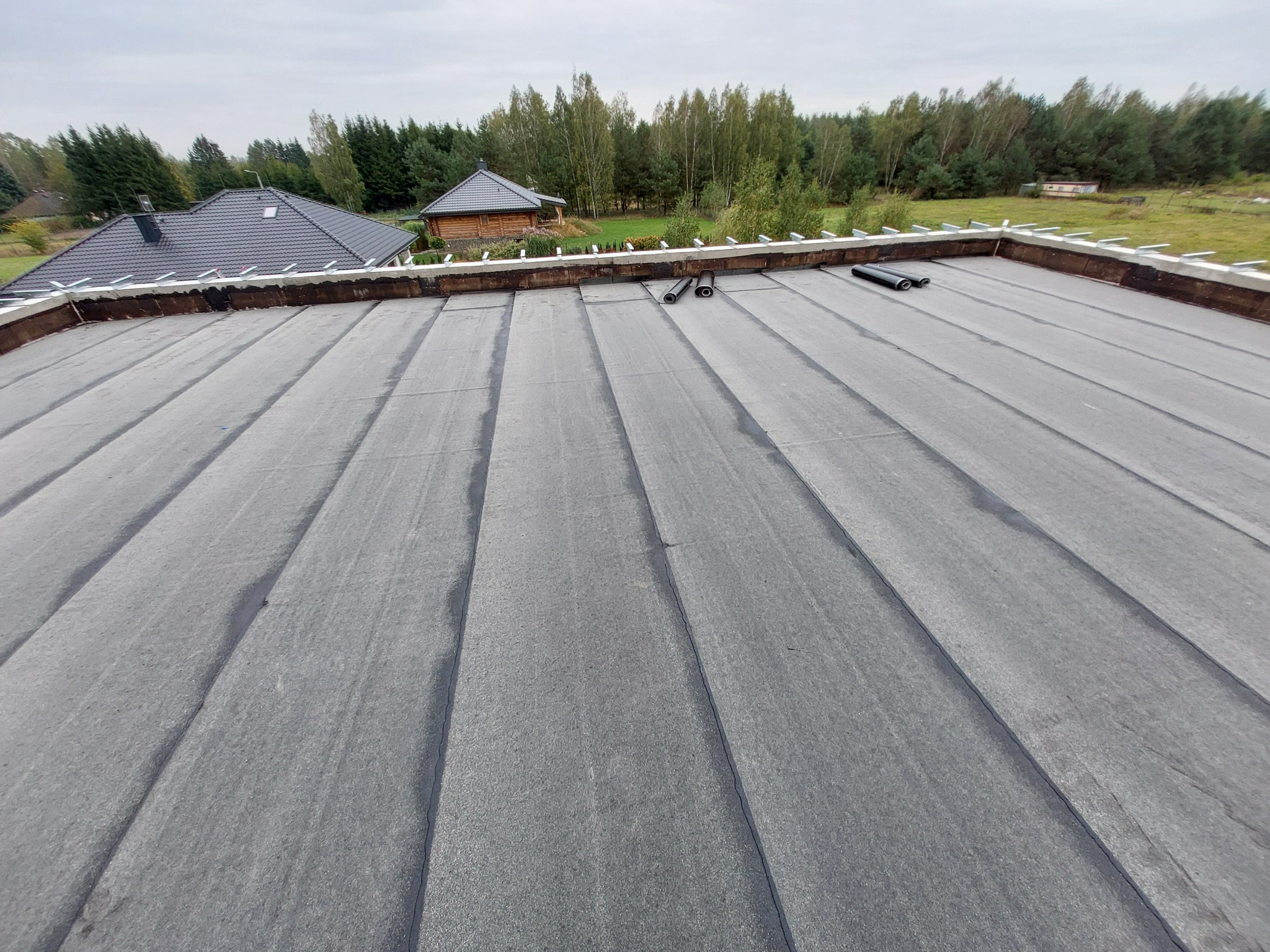 Szary dach pokryty papą termozgrzewalną z widocznymi rolkami papy i elementami konstrukcji dachu w tle, na tle zieleni i zabudowań.