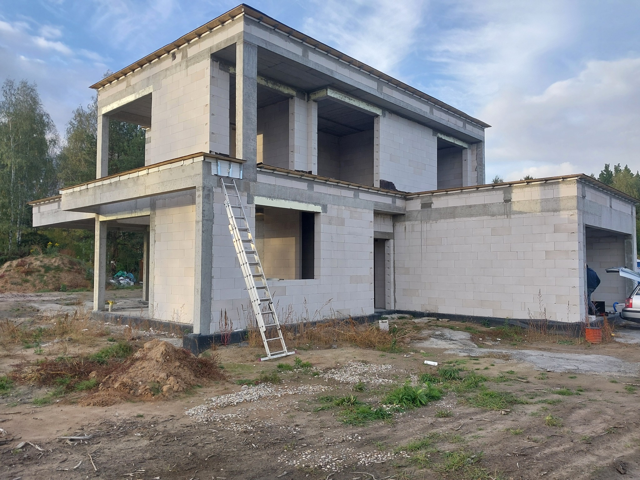 Surowy stan dwukondygnacyjnego domu z betonu komórkowego w trakcie budowy. Widoczna drabina oparta o ścianę i częściowo otwarty bagażnik samochodu w tle.