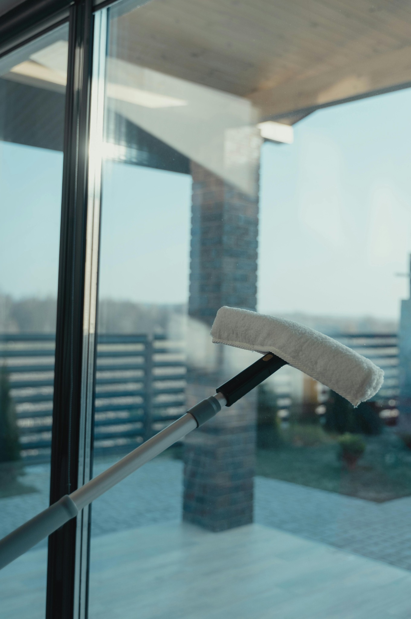 Mycie okna z zewnątrz za pomocą mopa na teleskopowej rączce. Widok na dom z ogrodem przez czyszczoną szybę.