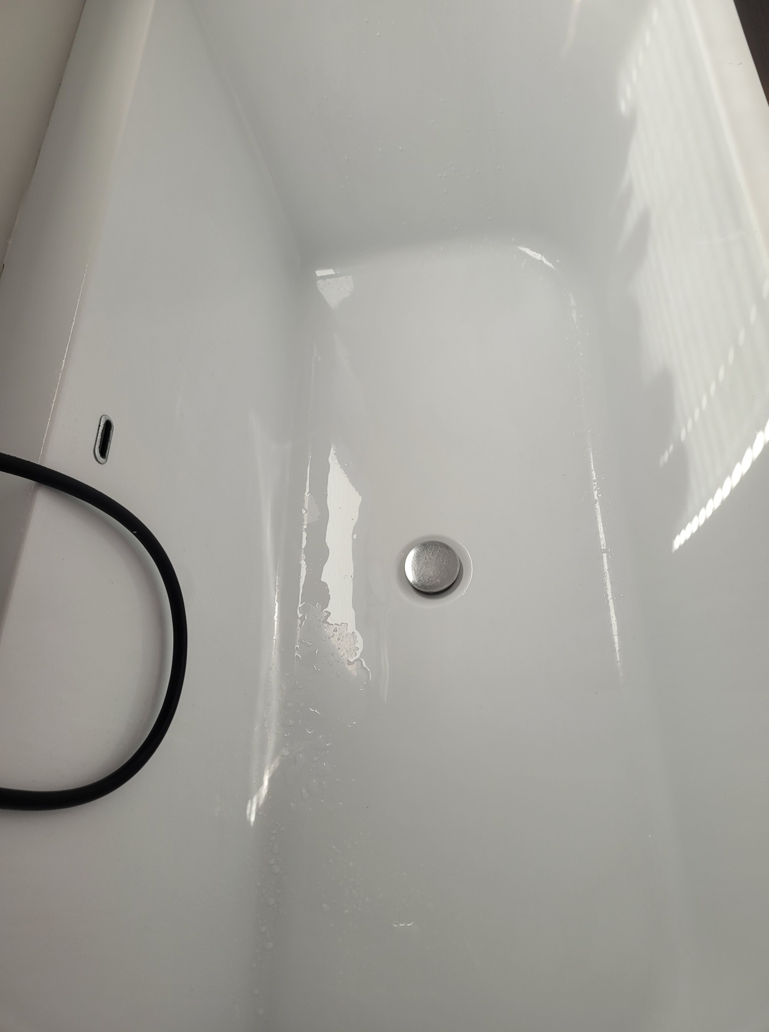 Biała, wyczyszczona wanna z odpływem, na której widoczne są resztki wody. Fragment czarnego węża w lewym dolnym rogu. Refleksy światła na powierzchni.