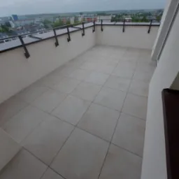 Jasne płytki na balkonie z metalową balustradą, widok na miasto w pochmurny dzień. Minimalistyczny design i symetryczny układ płytek podnoszą estetykę przestrzeni.