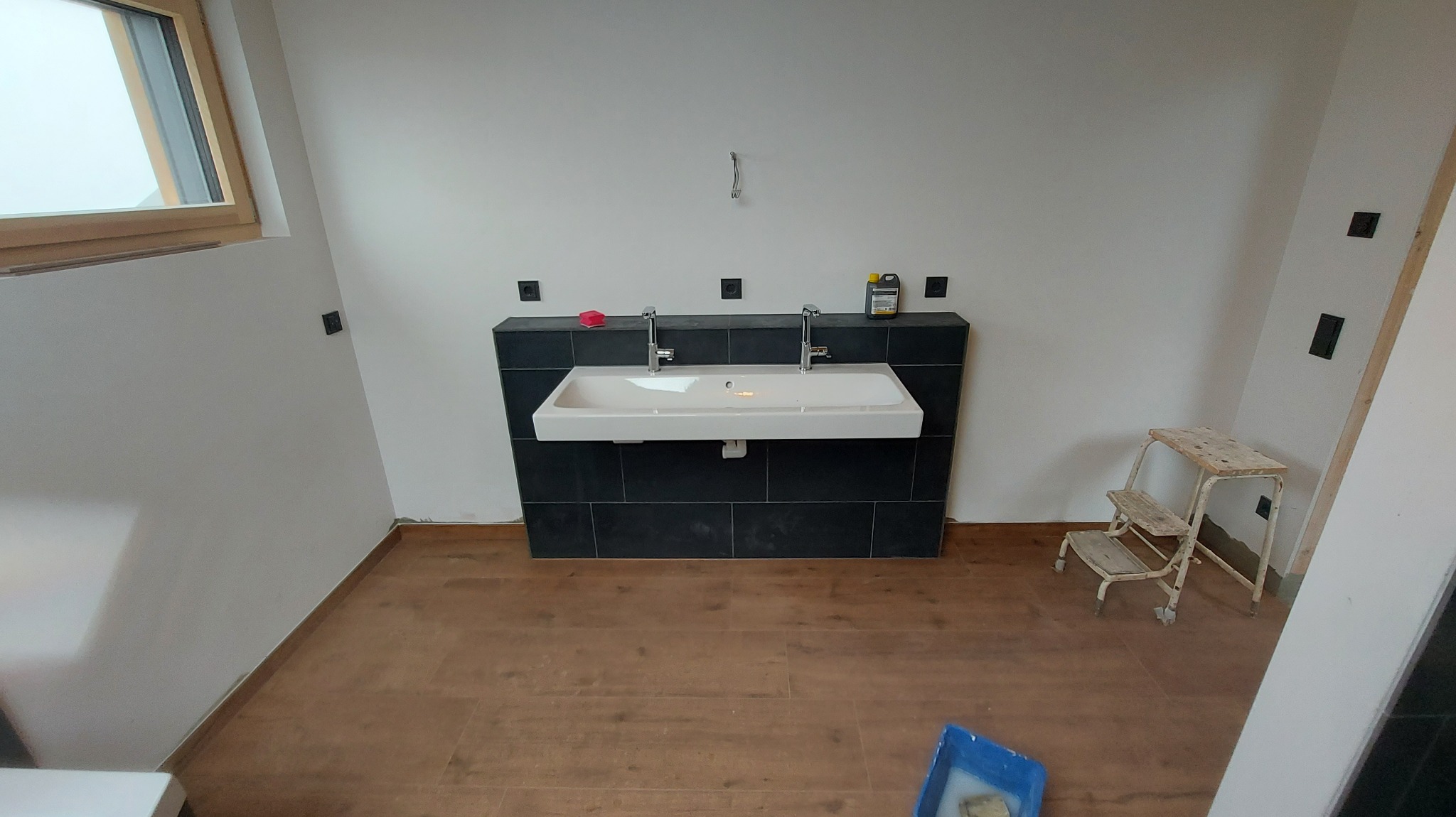 Łazienka w trakcie wykańczania: podwójna umywalka z czarną glazurą, drewnopodobna podłoga, drabinka malarska i okno. Widoczne puszki elektryczne.