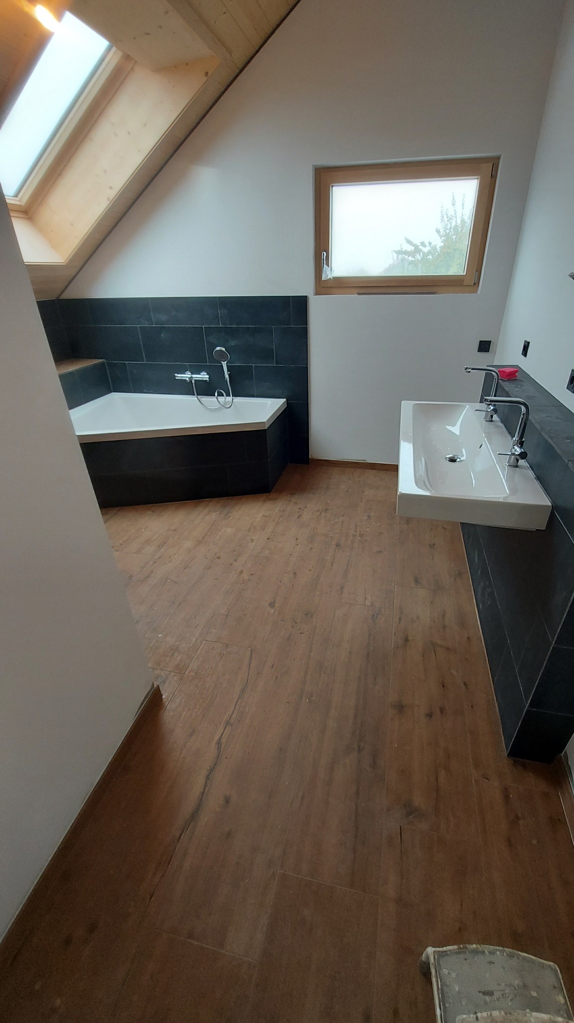 Jasna łazienka na poddaszu z drewnopodobną podłogą, czarnymi płytkami przy wannie i umywalce. Okno dachowe i zwykłe okno. Minimalistyczny styl.