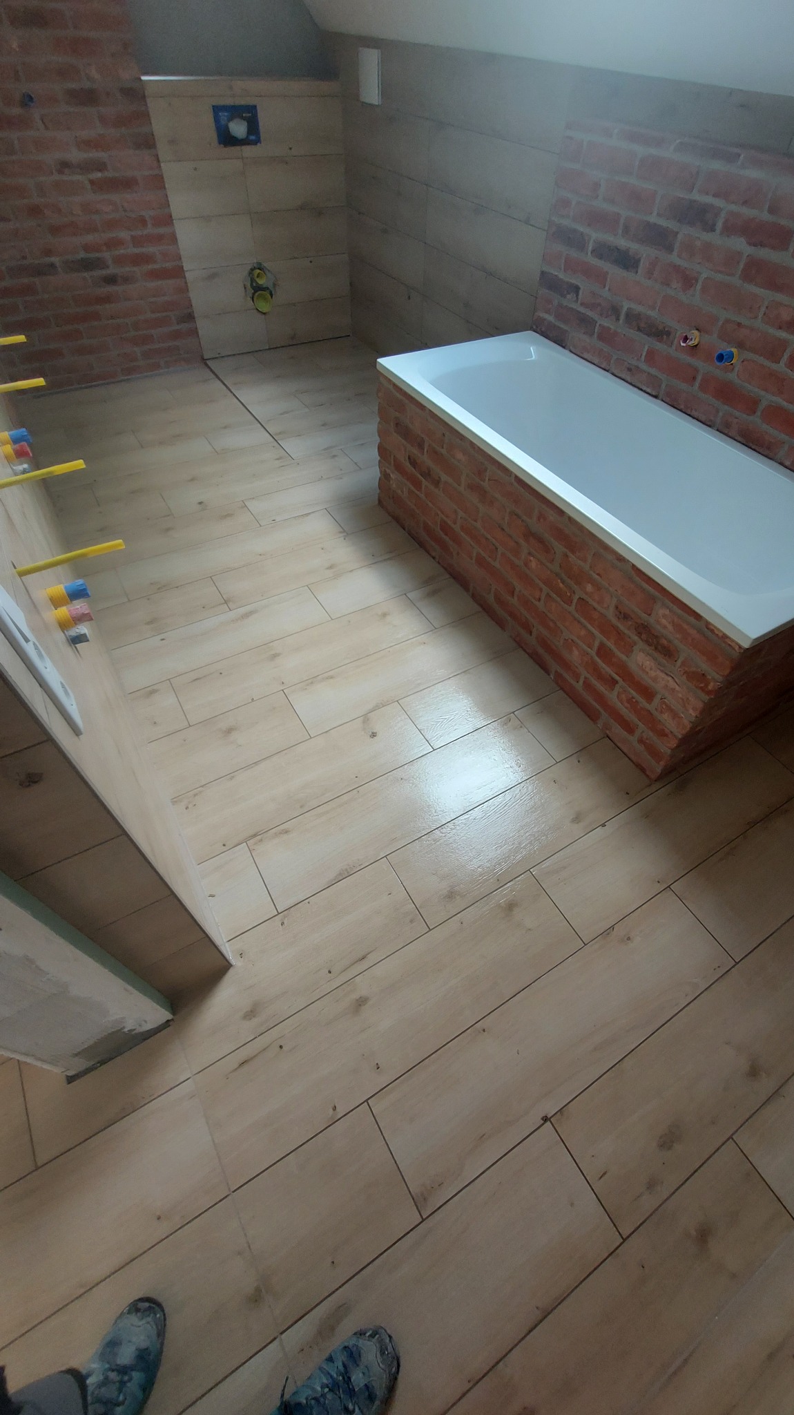 Ukończona łazienka na poddaszu z drewnopodobną podłogą, obudową wanny z cegły i częściowo wykończonymi ścianami. Widoczne narzędzia glazurnicze.