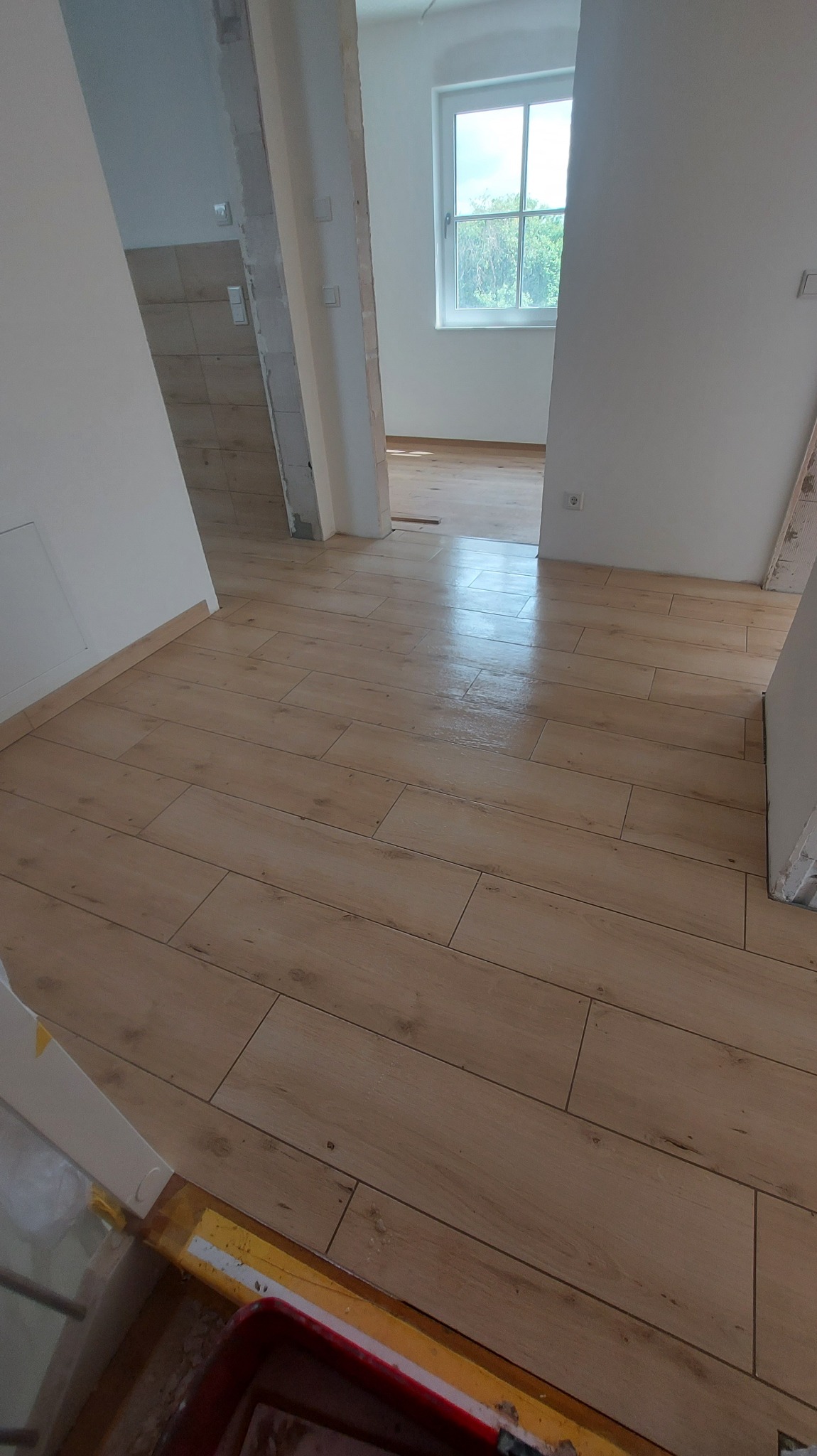 Ukończona podłoga z płytek imitujących drewno w jasnym wnętrzu, widok na okno i sąsiednie pomieszczenia w trakcie remontu.