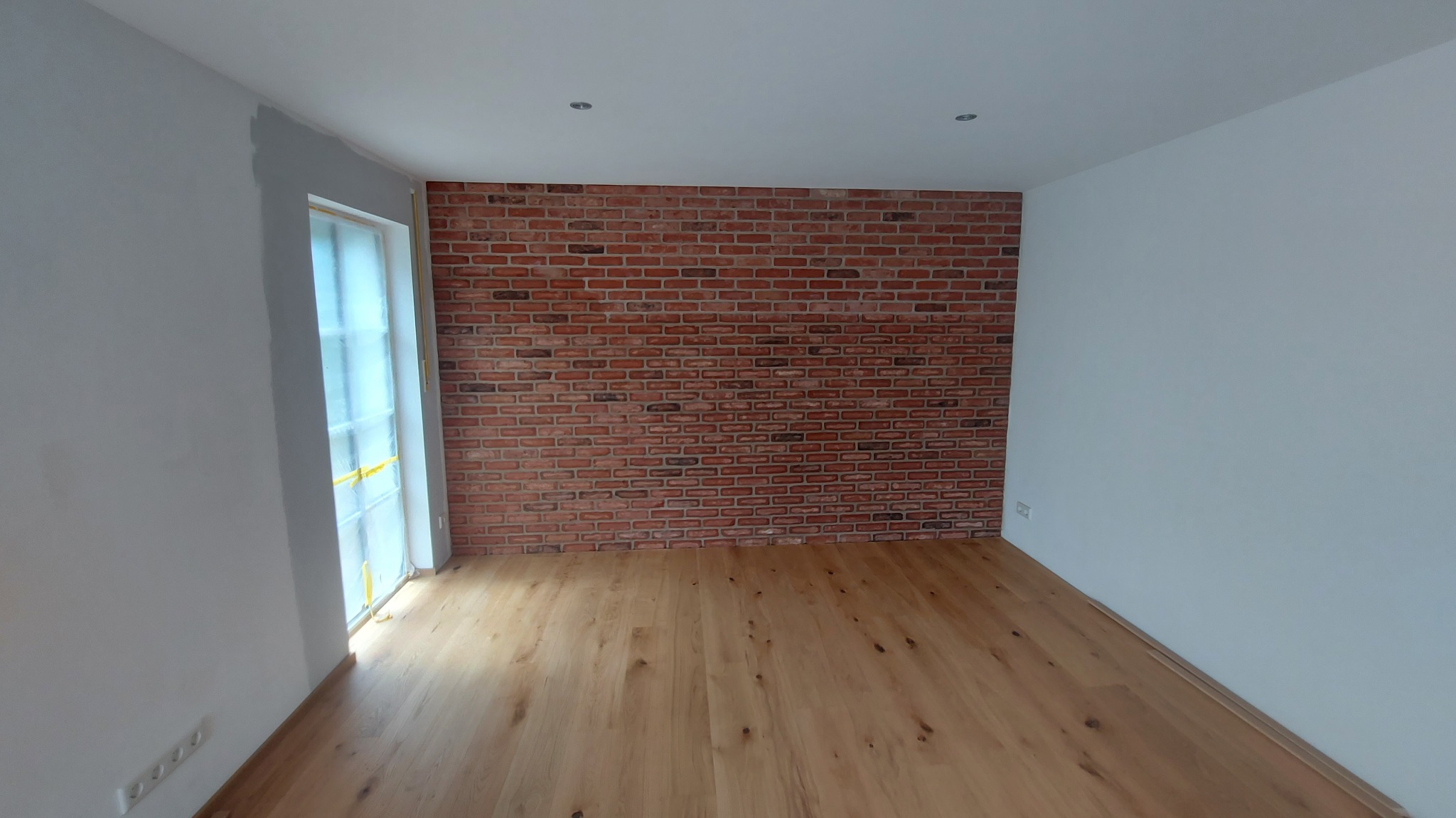 Pokój po remoncie: parkiet, ściana z cegły dekoracyjnej i białe ściany. Widoczne gniazdka elektryczne i okno zabezpieczone folią malarską.