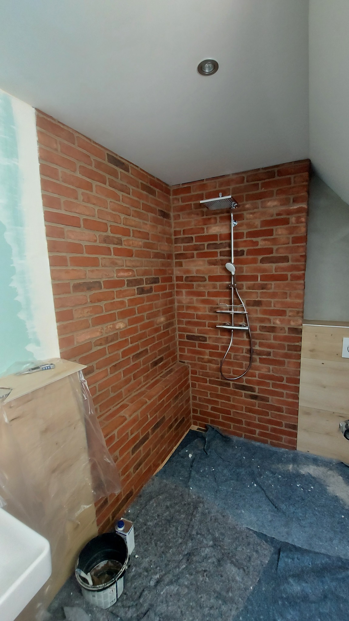 Wykończona kabina prysznicowa z ceglaną ścianą i chromowaną armaturą. Podłoga zabezpieczona folią ochronną. Widoczny fragment umywalki i ściany w trakcie przygotowania.