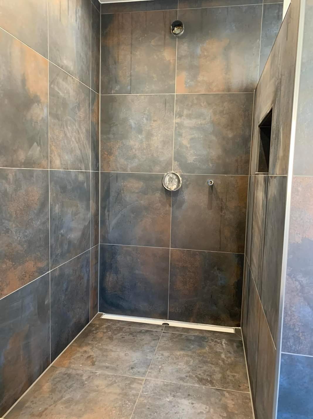 Stylowy prysznic z ciemnymi, metalicznymi płytkami, widoczny odpływ liniowy i otwór na baterię. Minimalistyczny design z akcentem na fakturę materiałów.