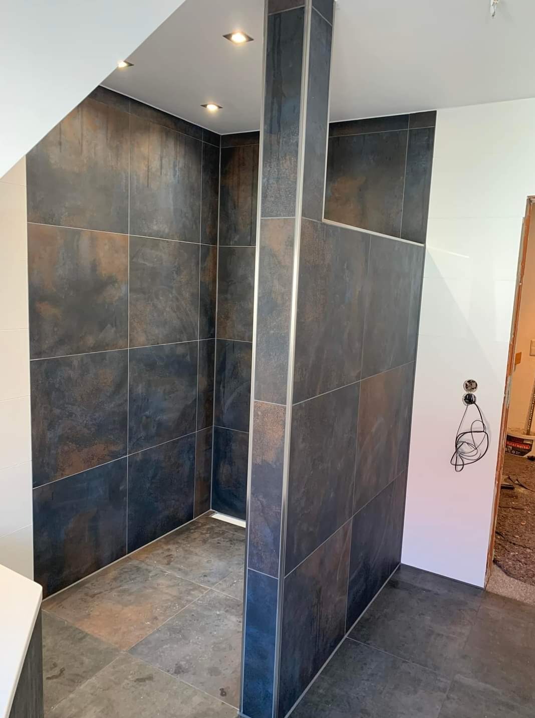 Nowoczesna kabina prysznicowa z ciemnymi, metalizowanymi płytkami i srebrnymi listwami wykończeniowymi. Widoczne oświetlenie punktowe i fragment niedokończonej ściany.