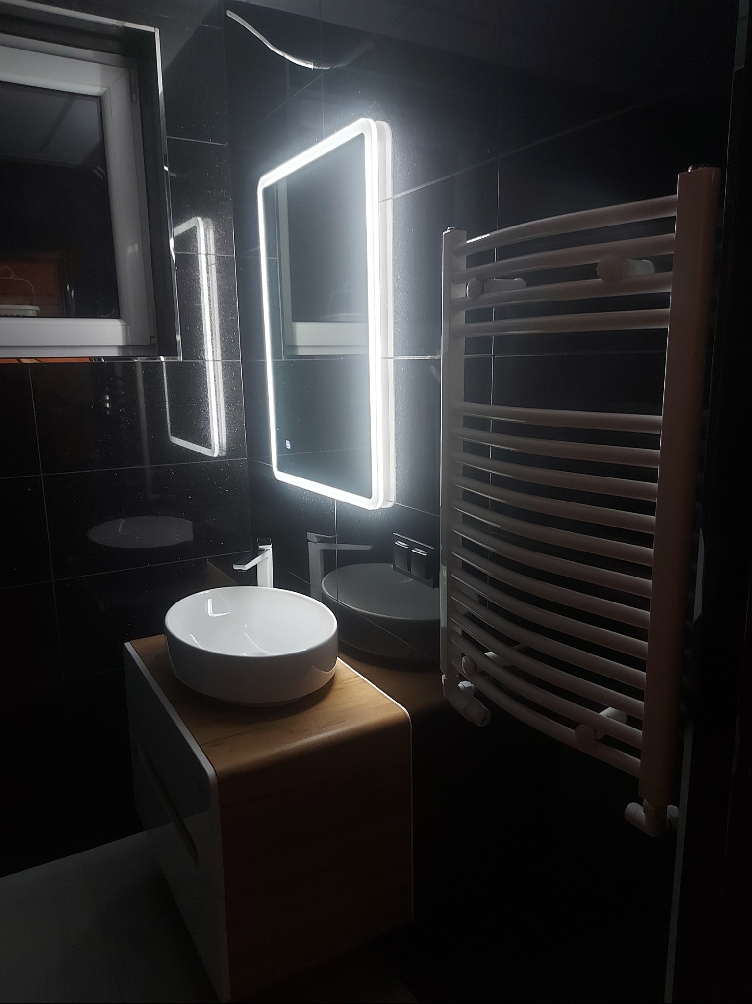 Nowoczesna łazienka z czarnymi płytkami, okrągłą umywalką na drewnianej szafce, podświetlanym lustrem i białym grzejnikiem drabinkowym.