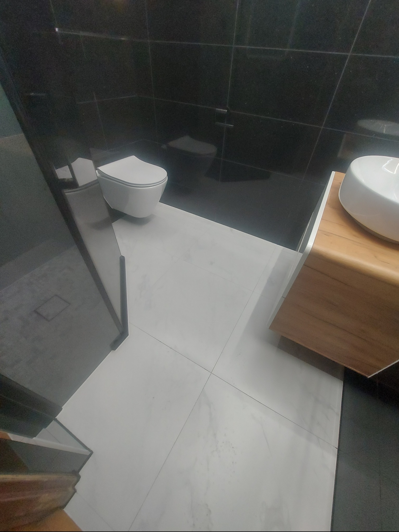 Nowoczesna łazienka z czarnymi płytkami na ścianach i białymi na podłodze. Umywalka na drewnianej szafce, wisząca toaleta. Minimalistyczny design.