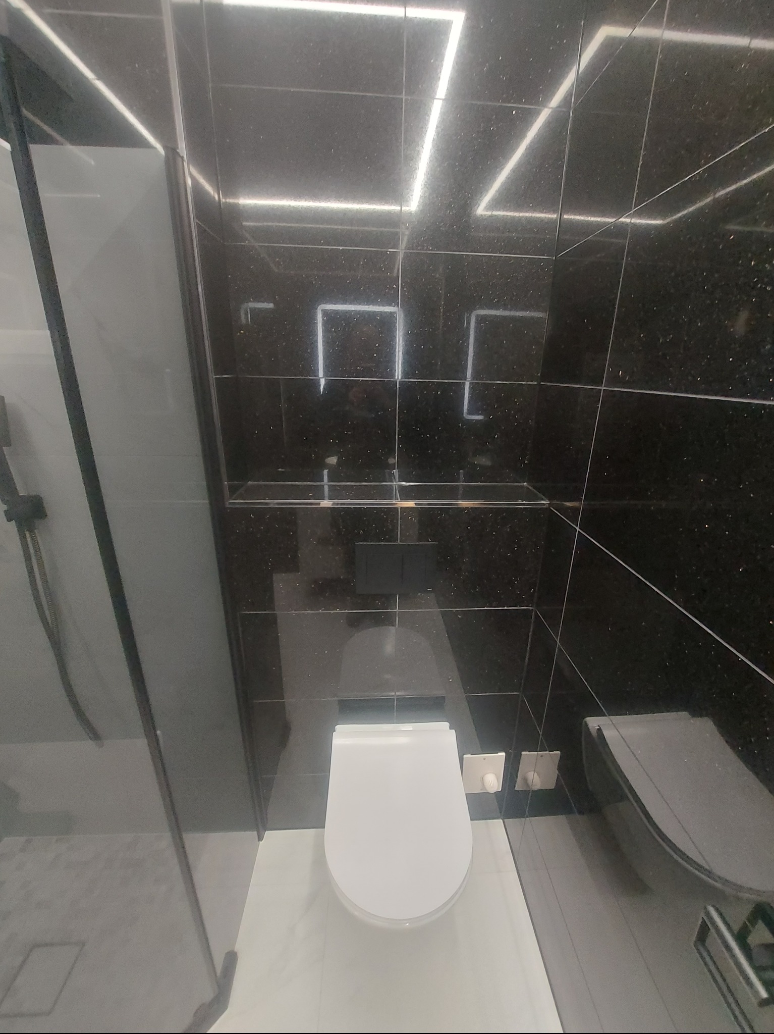 Nowoczesna łazienka z czarnymi, błyszczącymi płytkami imitującymi kamień, podświetleniem LED i minimalistyczną toaletą. Widok z kabiny prysznicowej.