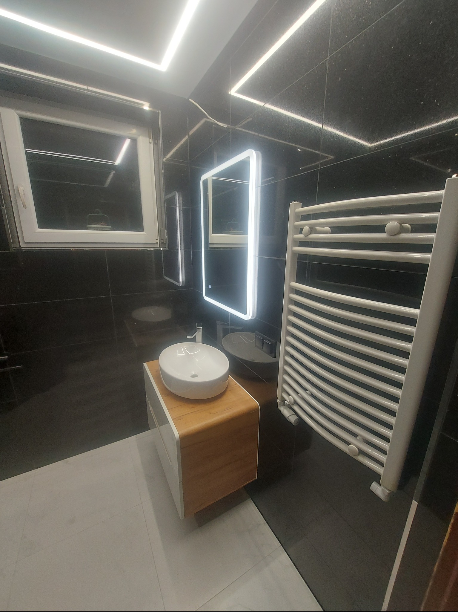 Nowoczesna łazienka z czarnymi płytkami, podświetlanym lustrem, białą umywalką nablatową na drewnianej szafce i grzejnikiem drabinkowym. Minimalistyczny design.