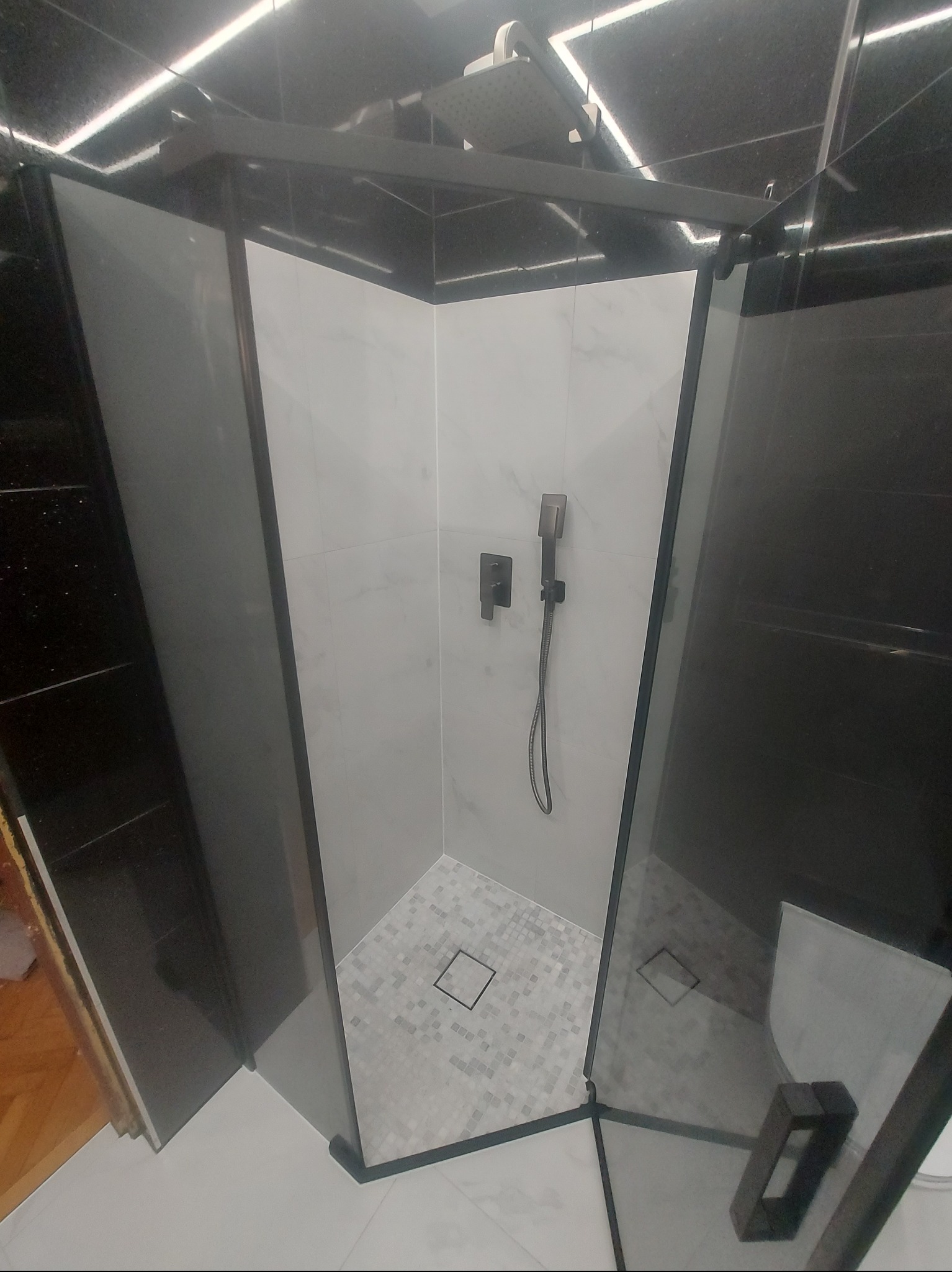 Nowoczesna kabina prysznicowa z czarnymi ramami i armaturą. Białe płytki imitujące marmur na ścianach, mozaika na podłodze. Widoczny odpływ liniowy i deszczownica.