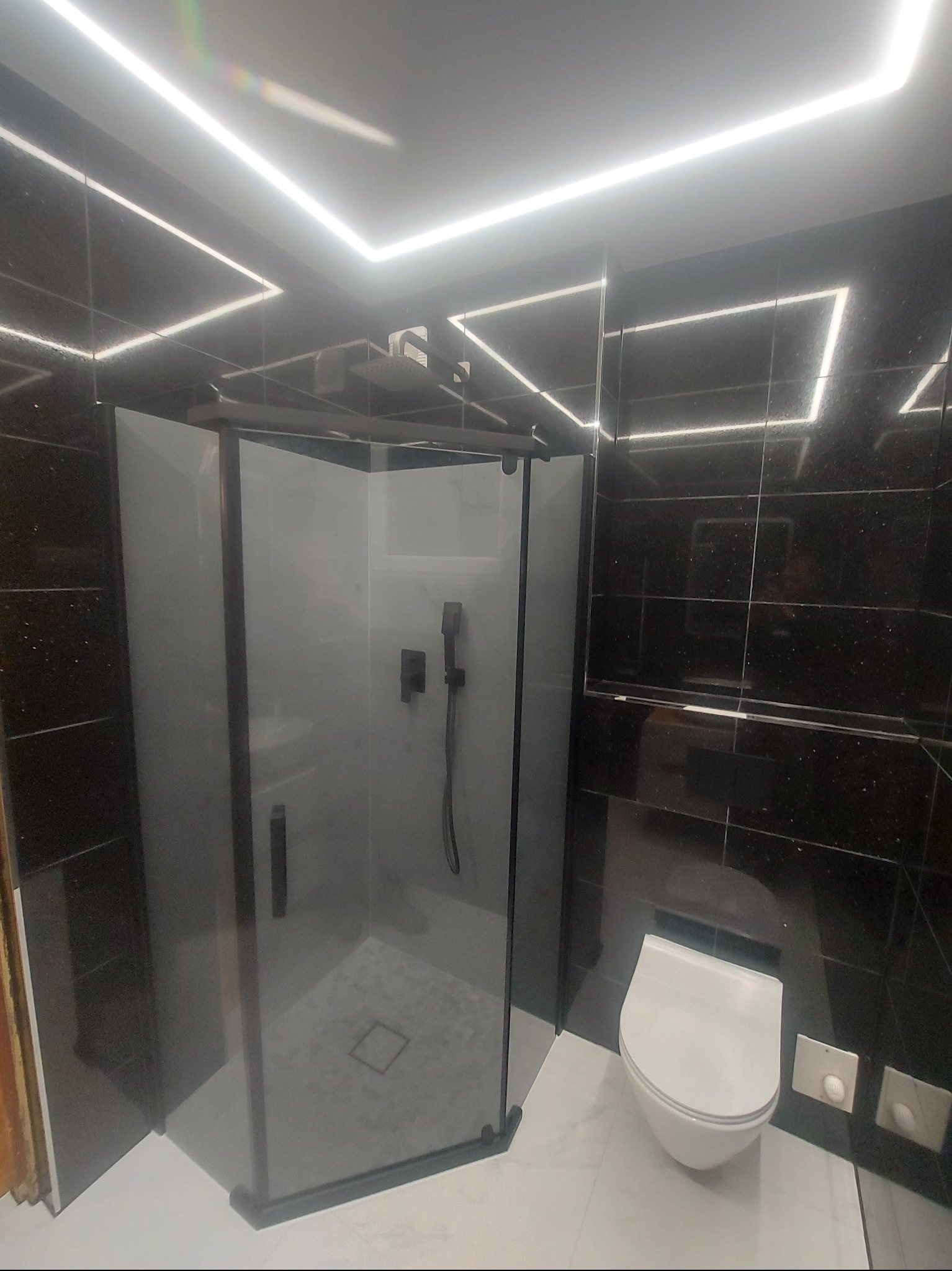 Nowoczesna łazienka z czarnymi, błyszczącymi płytkami, kabiną prysznicową typu walk-in z czarnymi profilami i podświetleniem LED na suficie.