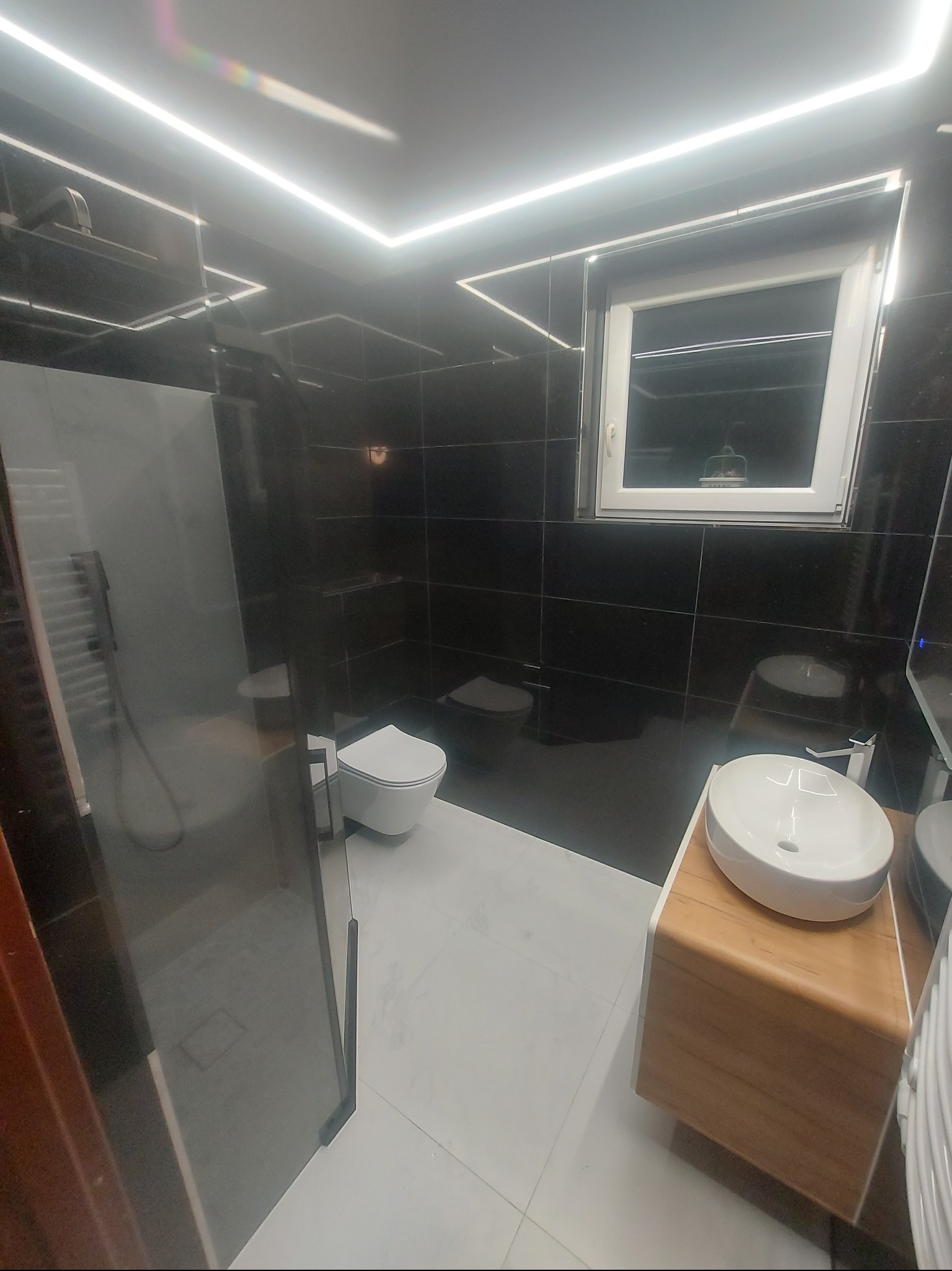 Nowoczesna łazienka z czarnymi płytkami, białymi elementami sanitarnymi i oświetleniem LED. Kabina prysznicowa ze szklanymi drzwiami, umywalka na drewnianej szafce.