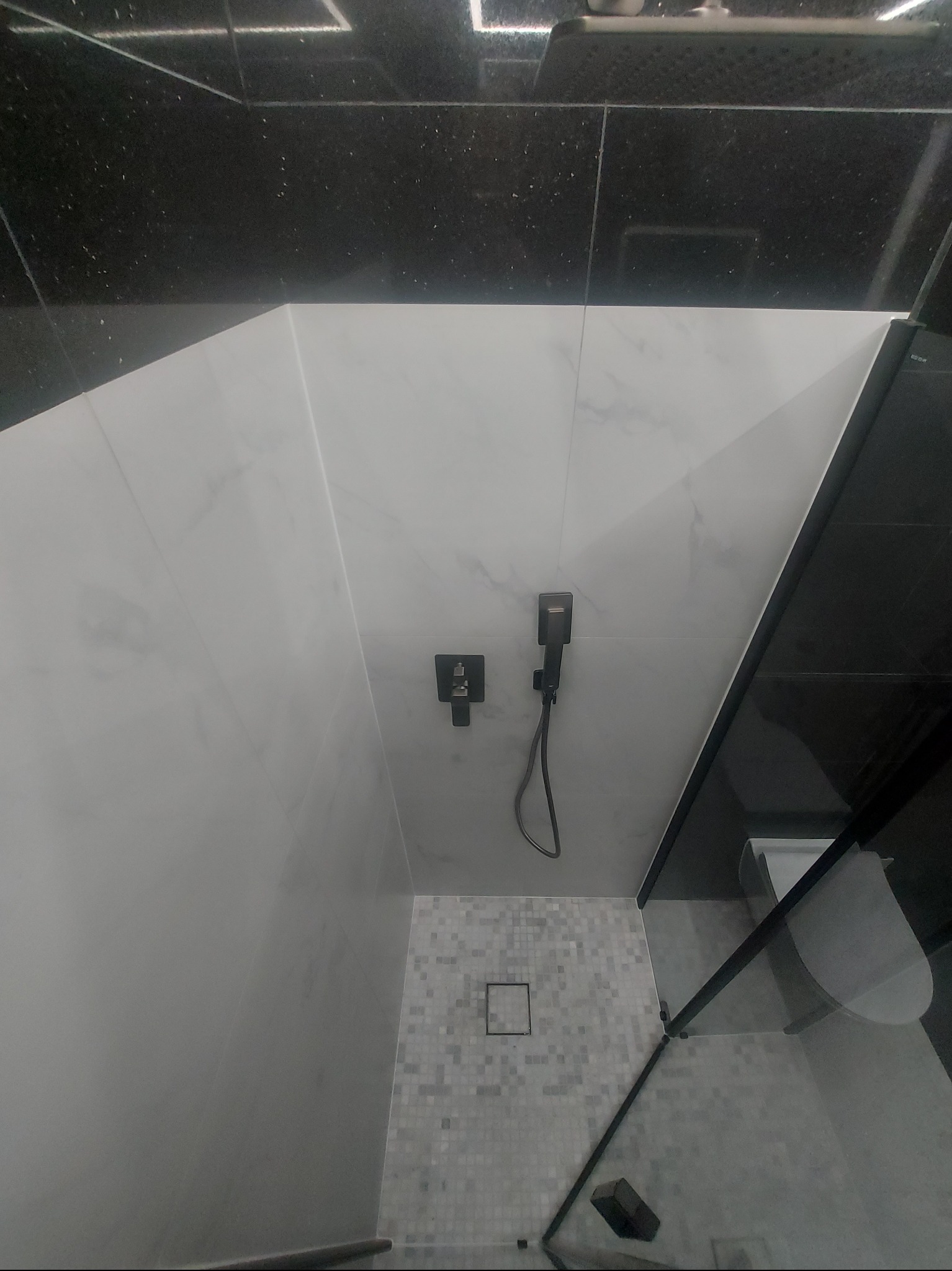 Nowoczesna kabina prysznicowa z czarną armaturą i białymi płytkami imitującymi marmur. Widoczny odpływ liniowy i szklane drzwi kabiny.