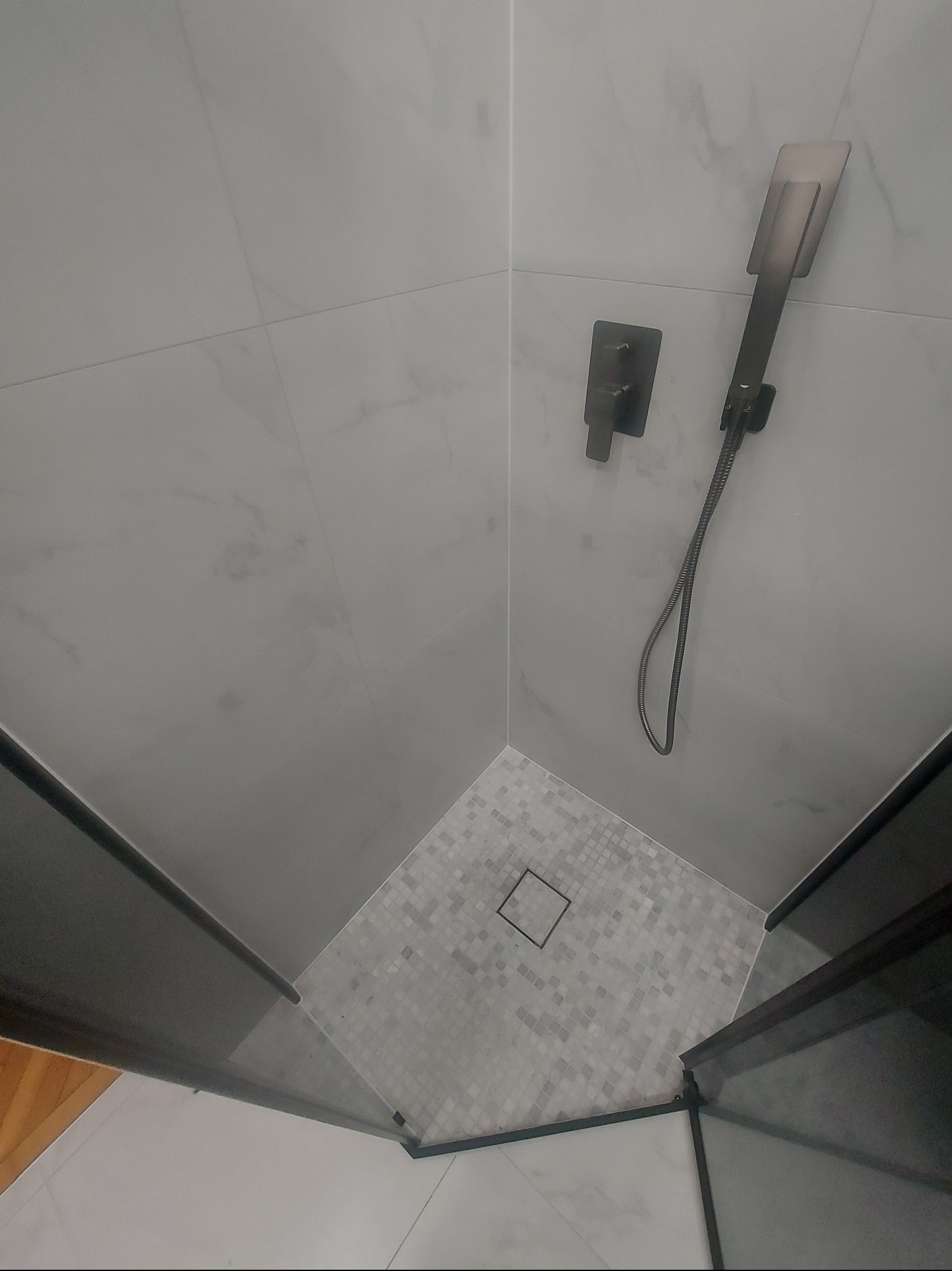 Nowoczesna kabina prysznicowa z szarymi płytkami imitującymi marmur na ścianach i mozaiką na podłodze. Czarna armatura i odpływ liniowy. Minimalistyczny design.