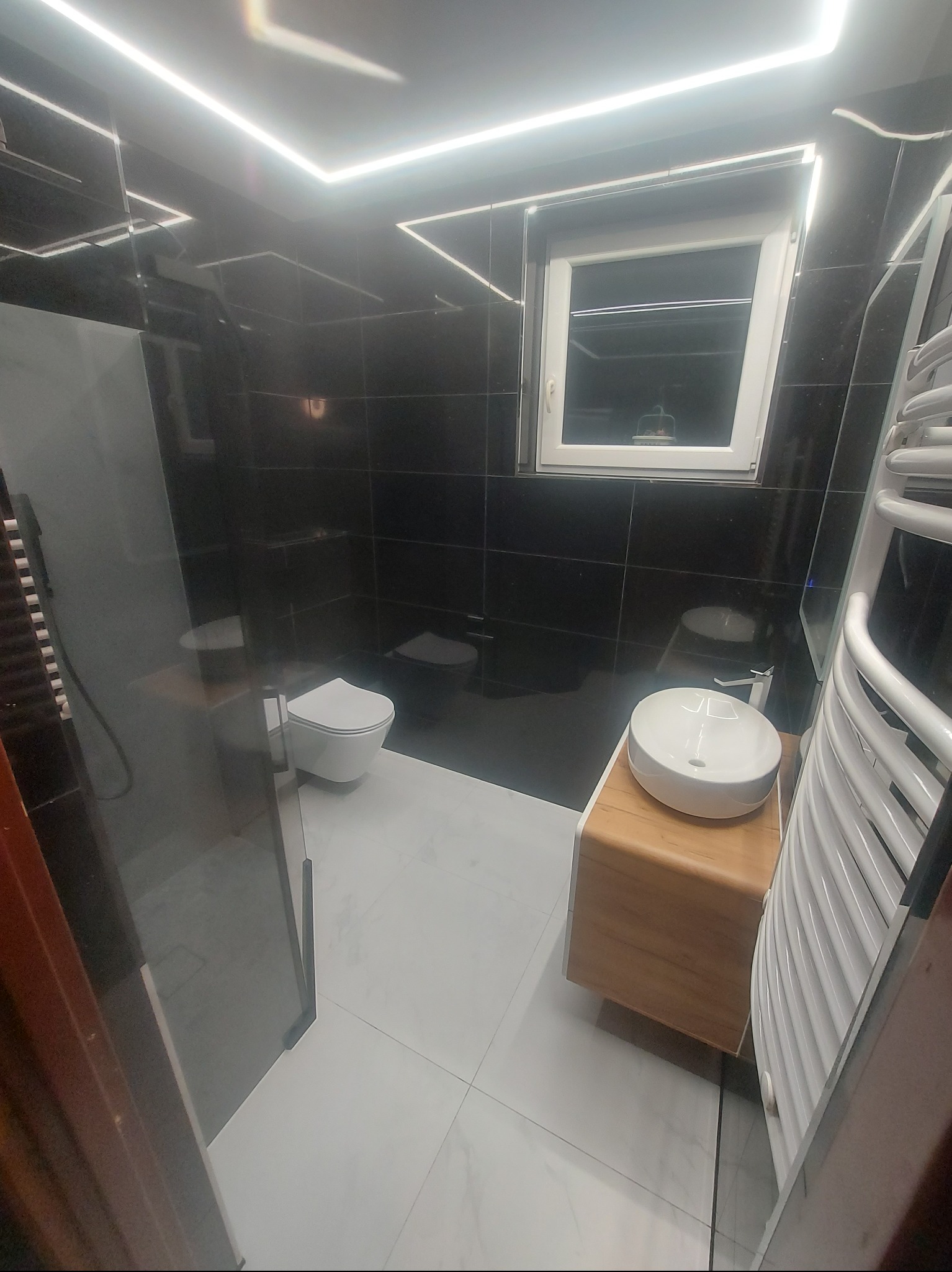 Nowoczesna łazienka z czarnymi płytkami, białymi sanitariatami i podświetleniem LED. Umywalka nablatowa na drewnianej szafce, grzejnik drabinkowy.