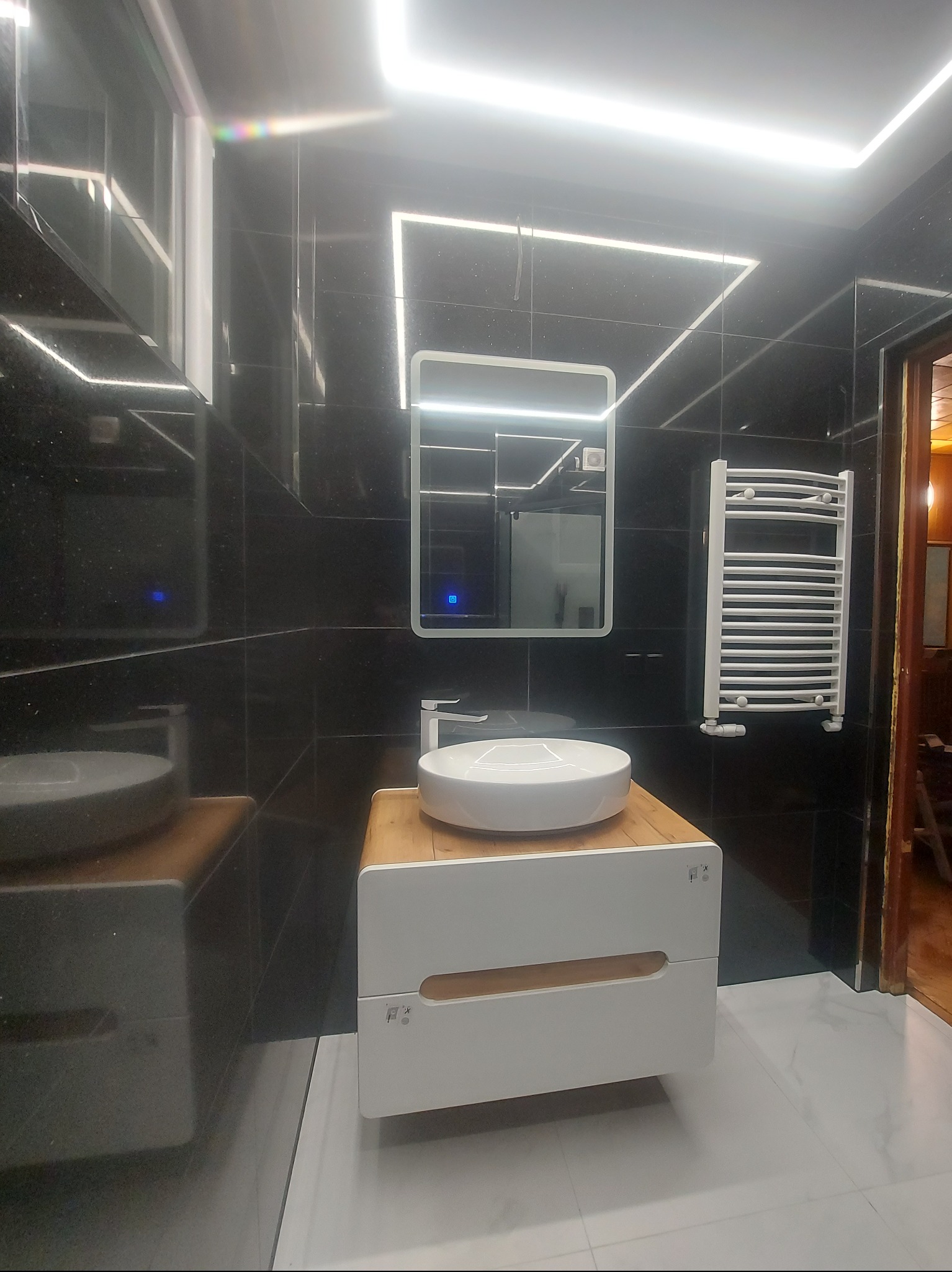 Nowoczesna łazienka z czarnymi płytkami, podświetlanym lustrem i białą umywalką nablatową na szafce z drewnianym akcentem. Grzejnik drabinkowy i oświetlenie LED.