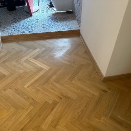 Michał Kozłowski - Parkiet ułożony w jodełkę w przejściu do łazienki z wzorzystymi płytkami. Jasne drewno, białe ściany, listwy przypodłogowe. Widok z góry, naturalne światło.