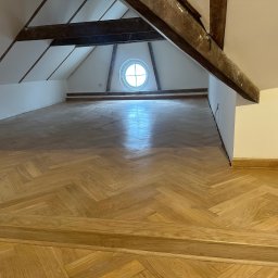 Michał Kozłowski - Ukończona podłoga z parkietu ułożonego w jodełkę w mansardzie w Warszawie. Widoczne drewniane belki stropowe i okrągłe okno.