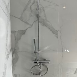 Michał Kozłowski - Bateria prysznicowa z deszczownicą na tle białych płytek imitujących marmur. Widoczne chromowane elementy i precyzyjne ułożenie płytek.