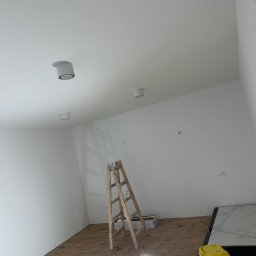 Michał Kozłowski - Wnętrze po malowaniu ścian na biało, widoczna drewniana podłoga, drabina malarska, lampa halogenowa i żółty odkurzacz. Oświetlenie sufitowe.