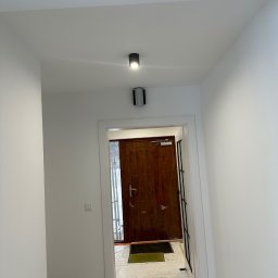 Michał Kozłowski - Biały korytarz po remoncie z drewnianymi drzwiami wejściowymi i nowoczesnym oświetleniem sufitowym. Widoczne zabezpieczenia podłogi.