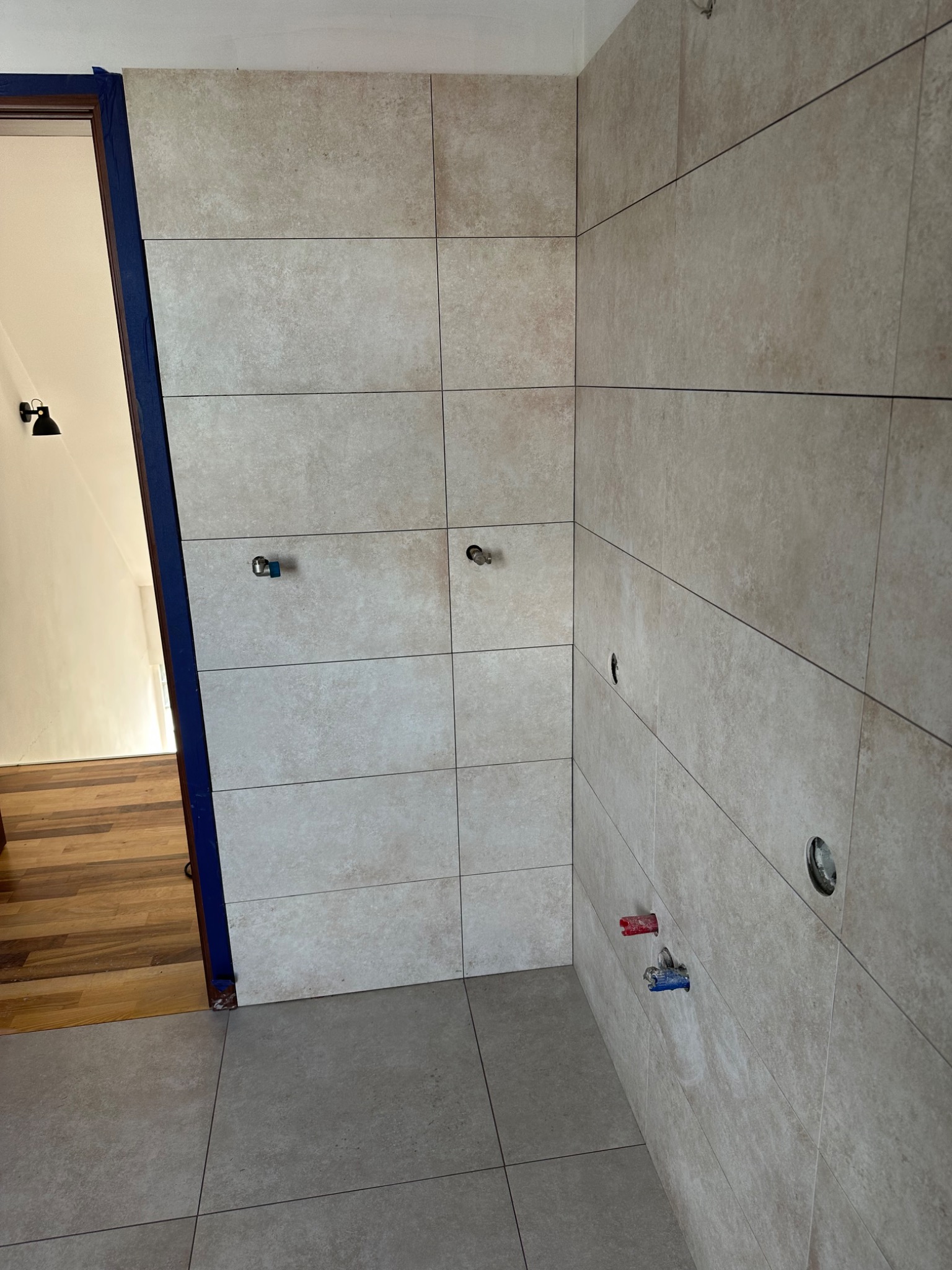 Wykończona łazienka z beżowymi, prostokątnymi płytkami. Widoczne wyprowadzenia wodne i otwory na armaturę. Drzwi z niebieskim zabezpieczeniem.