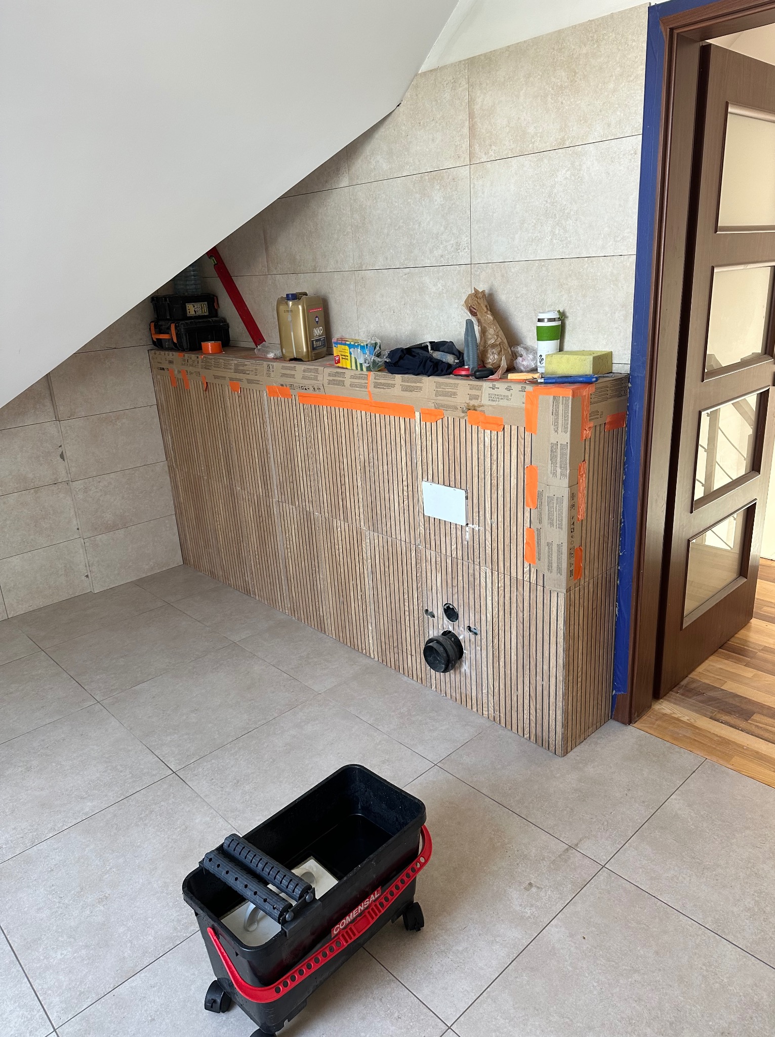 Łazienka w trakcie wykończenia: obudowa stelaża WC z pionowymi panelami imitującymi drewno, zabezpieczona kartonem i taśmą, narzędzia glazurnicze w wiadrze.