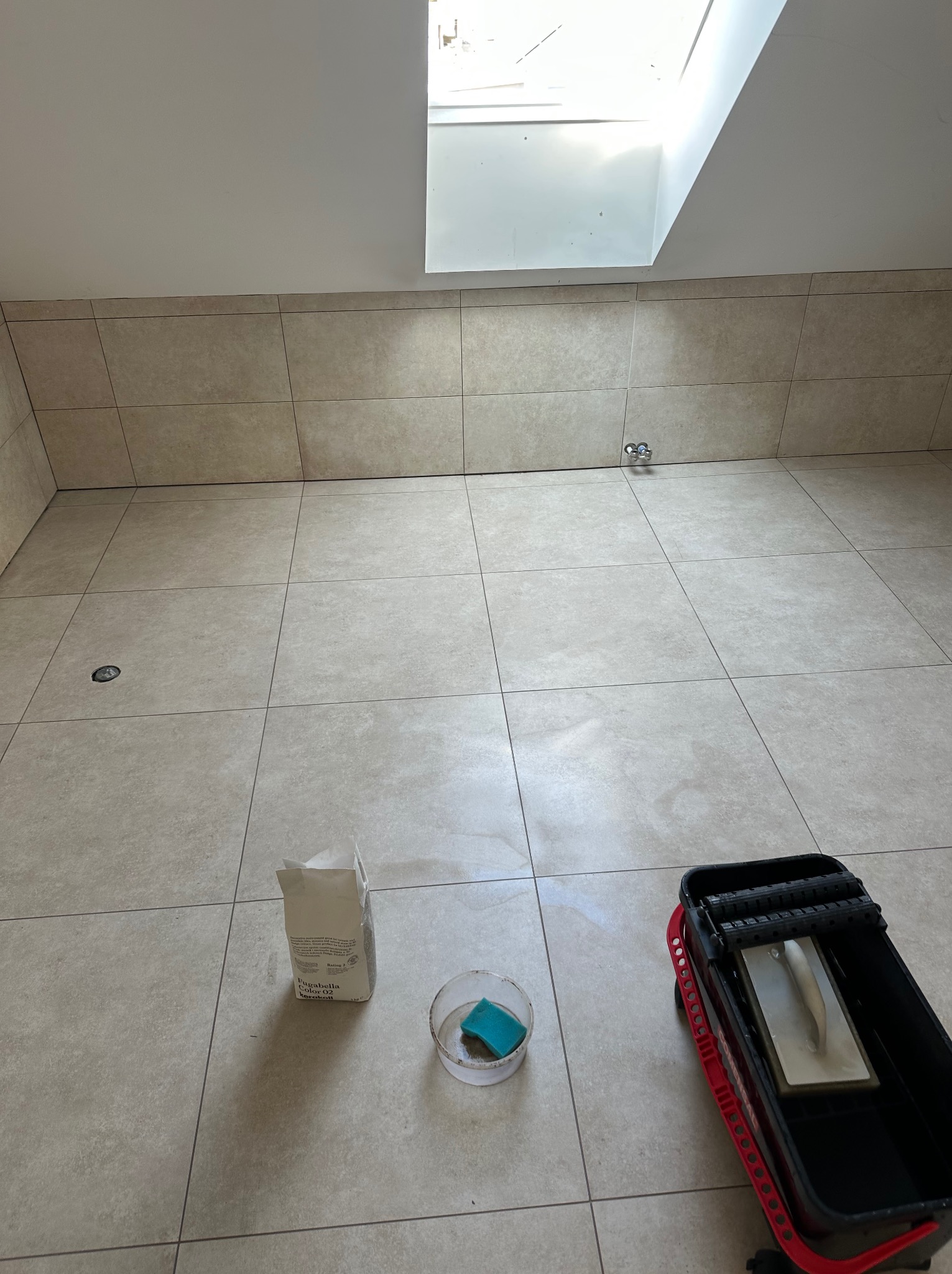 Wykończona podłoga w łazience z beżowych płytek. Widoczny worek z fugą, pojemnik z gąbką i wiadro do mycia płytek. Ujęcie z dołu, w tle okno dachowe.