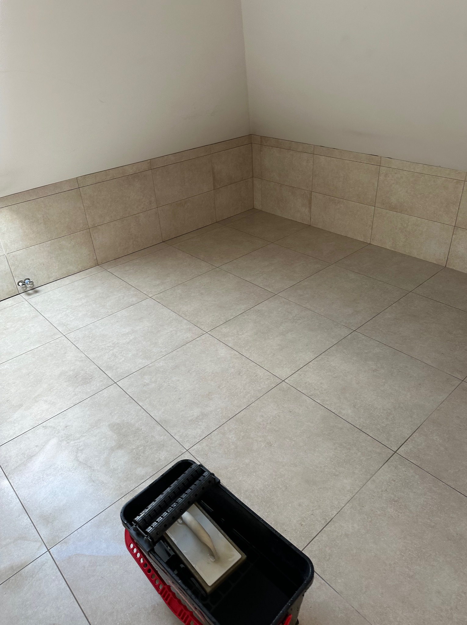 Wykończona beżowa podłoga i fragment ściany z płytek w łazience. W rogu wiadro glazurnicze. Widoczny kran. Minimalistyczny design.