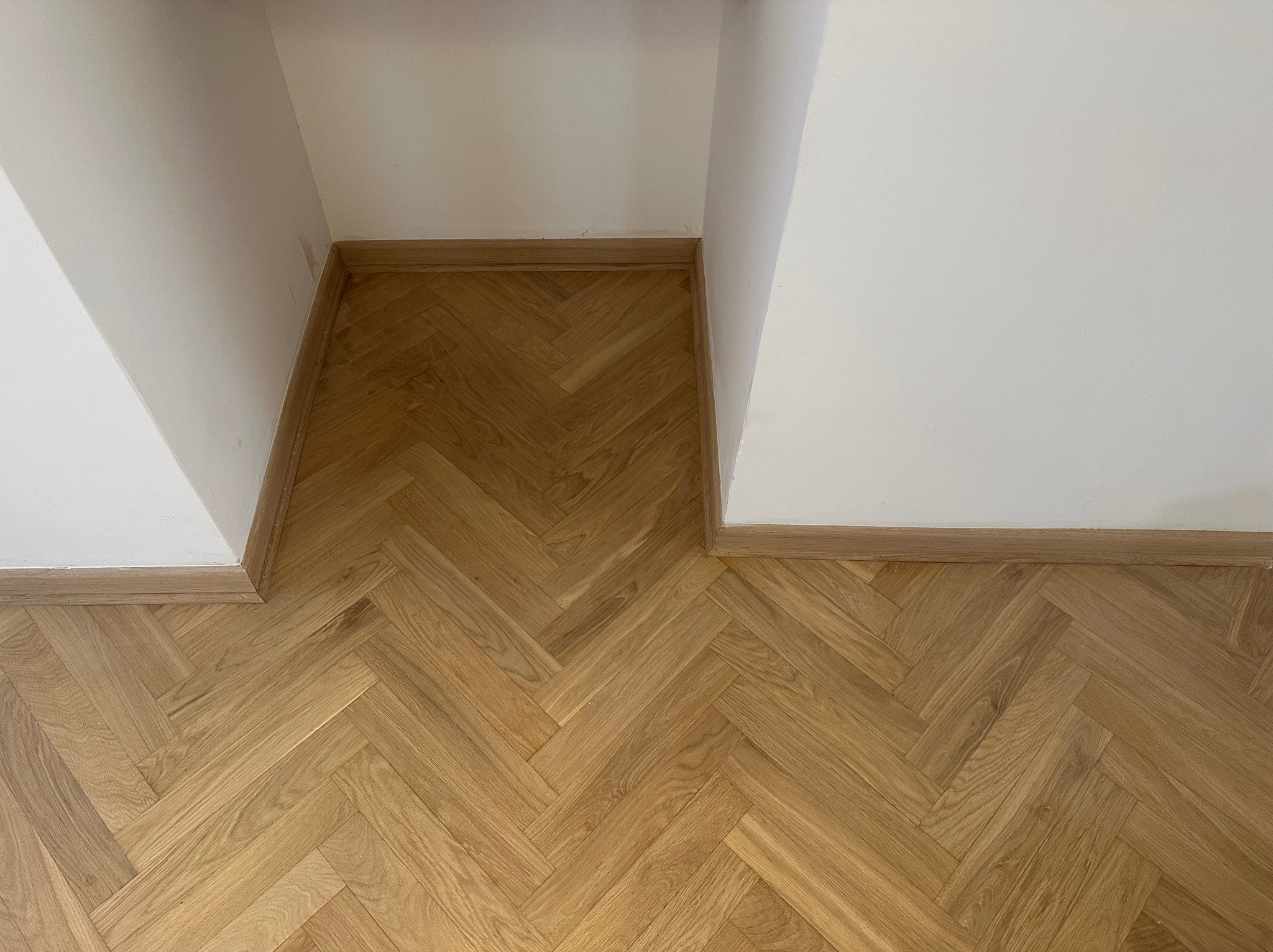 Parkiet ułożony we wzór jodełki francuskiej, listwy przypodłogowe w kolorze drewna, białe ściany. Widok z góry na fragment podłogi w rogu pomieszczenia.