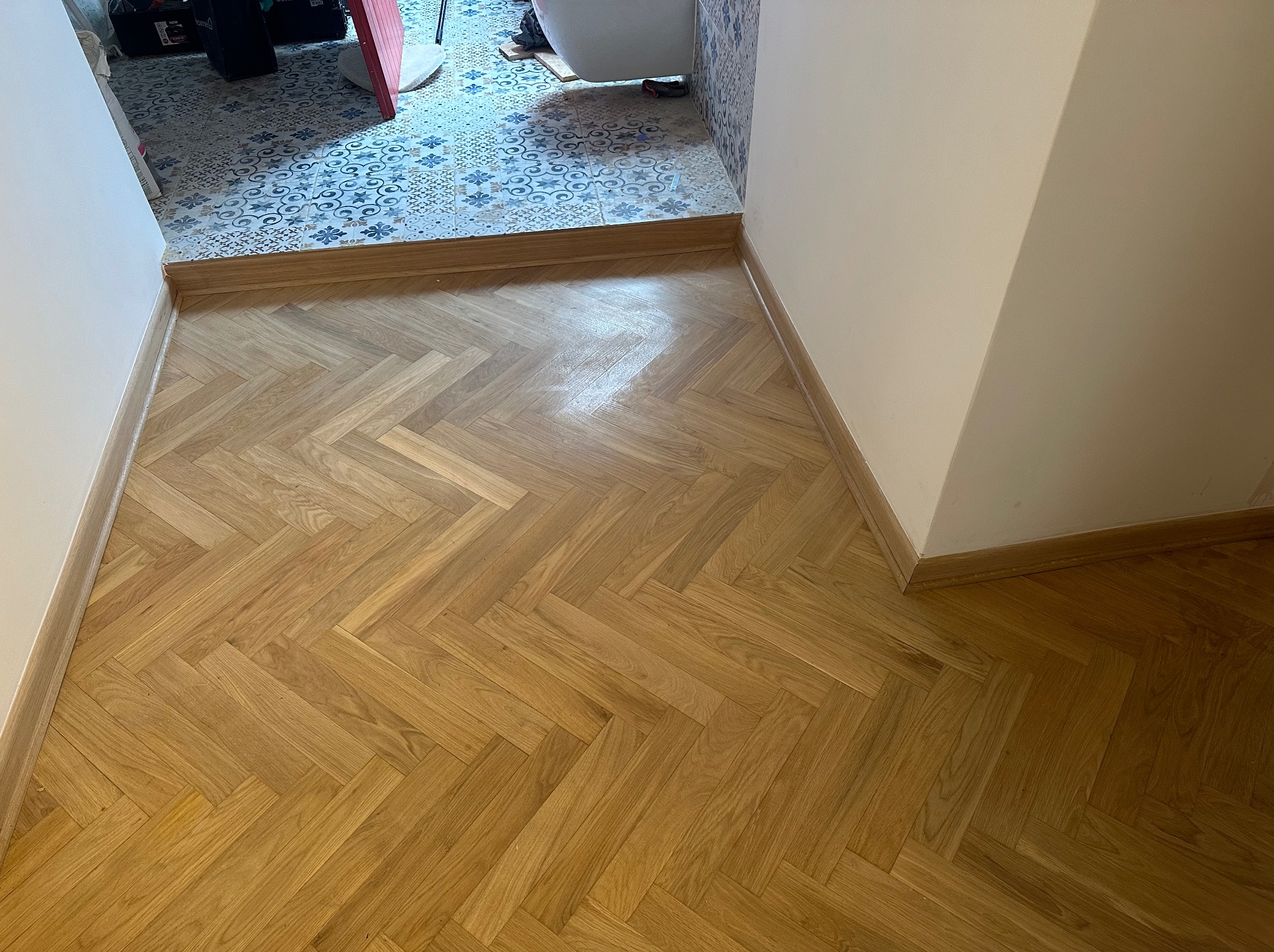 Parkiet ułożony w jodełkę w przejściu do łazienki z wzorzystymi płytkami. Jasne drewno, białe ściany, listwy przypodłogowe. Widok z góry, naturalne światło.