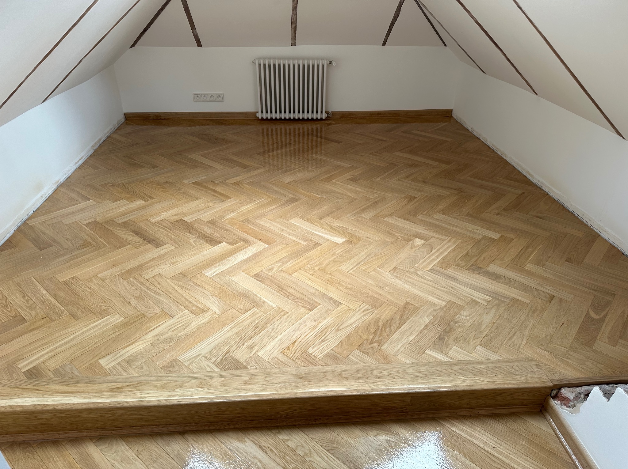 Parkiet w jodełkę po cyklinowaniu w jasnym pokoju na poddaszu. Widoczny grzejnik i listwy przypodłogowe. Charakterystyczne skosy sufitu.