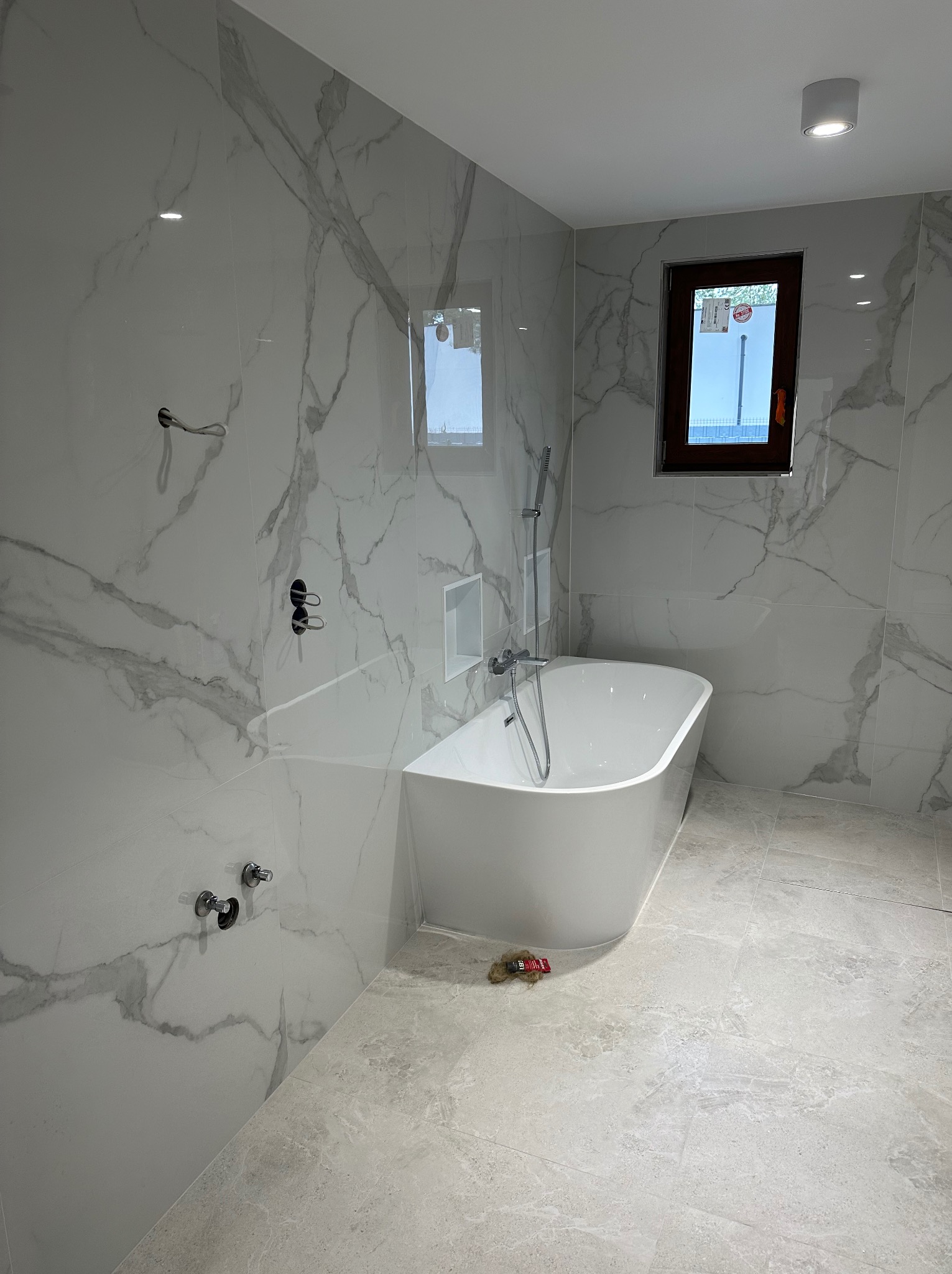 Nowoczesna łazienka: biała wanna wolnostojąca, marmurowe płytki ścienne i podłogowe, okno z brązową ramą. Minimalistyczny design, chromowana armatura.