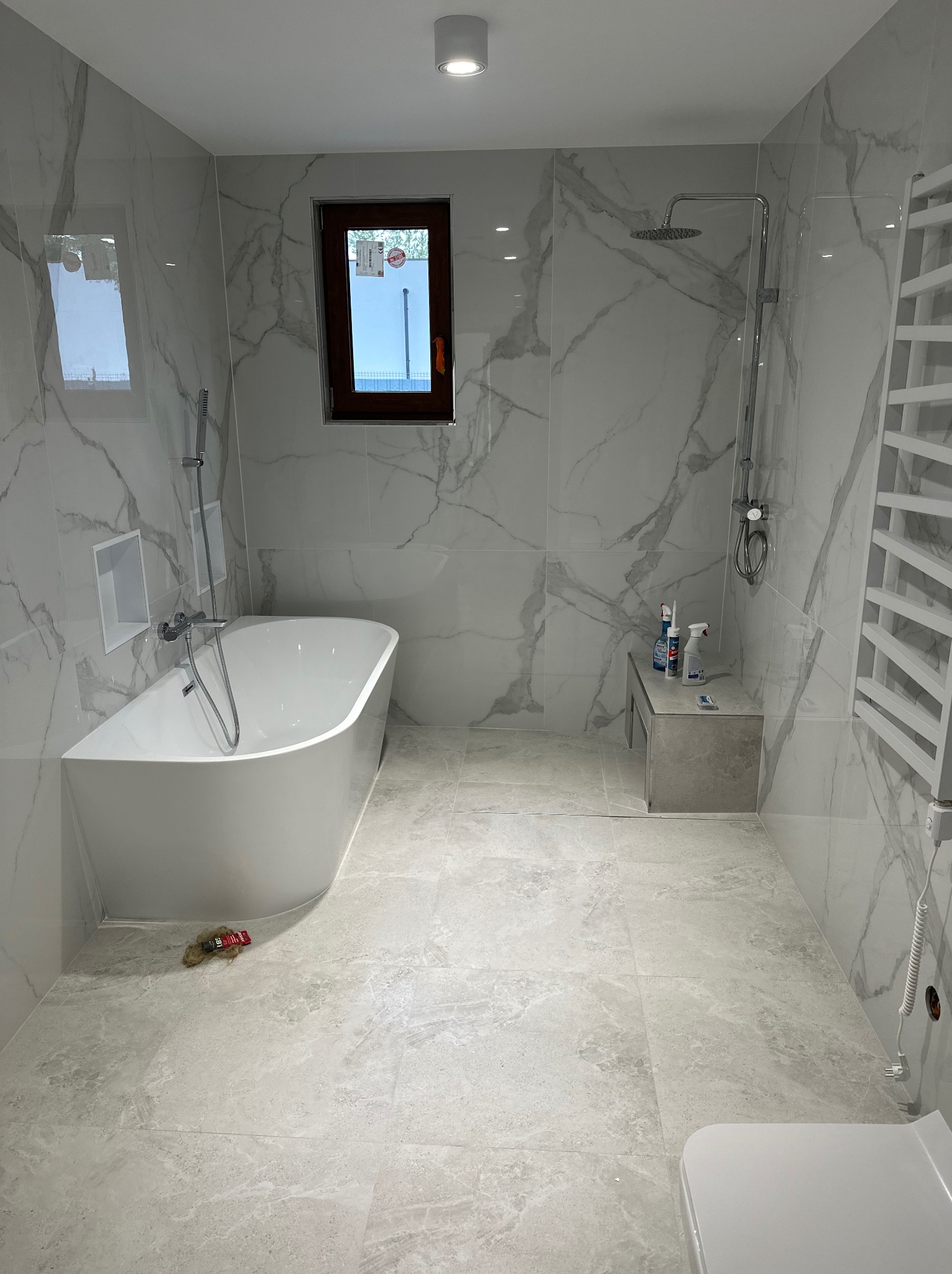 Nowoczesna łazienka z wolnostojącą wanną, marmurowymi płytkami i oknem z brązową ramą. Na podłodze widoczne resztki po montażu. Minimalistyczny design.