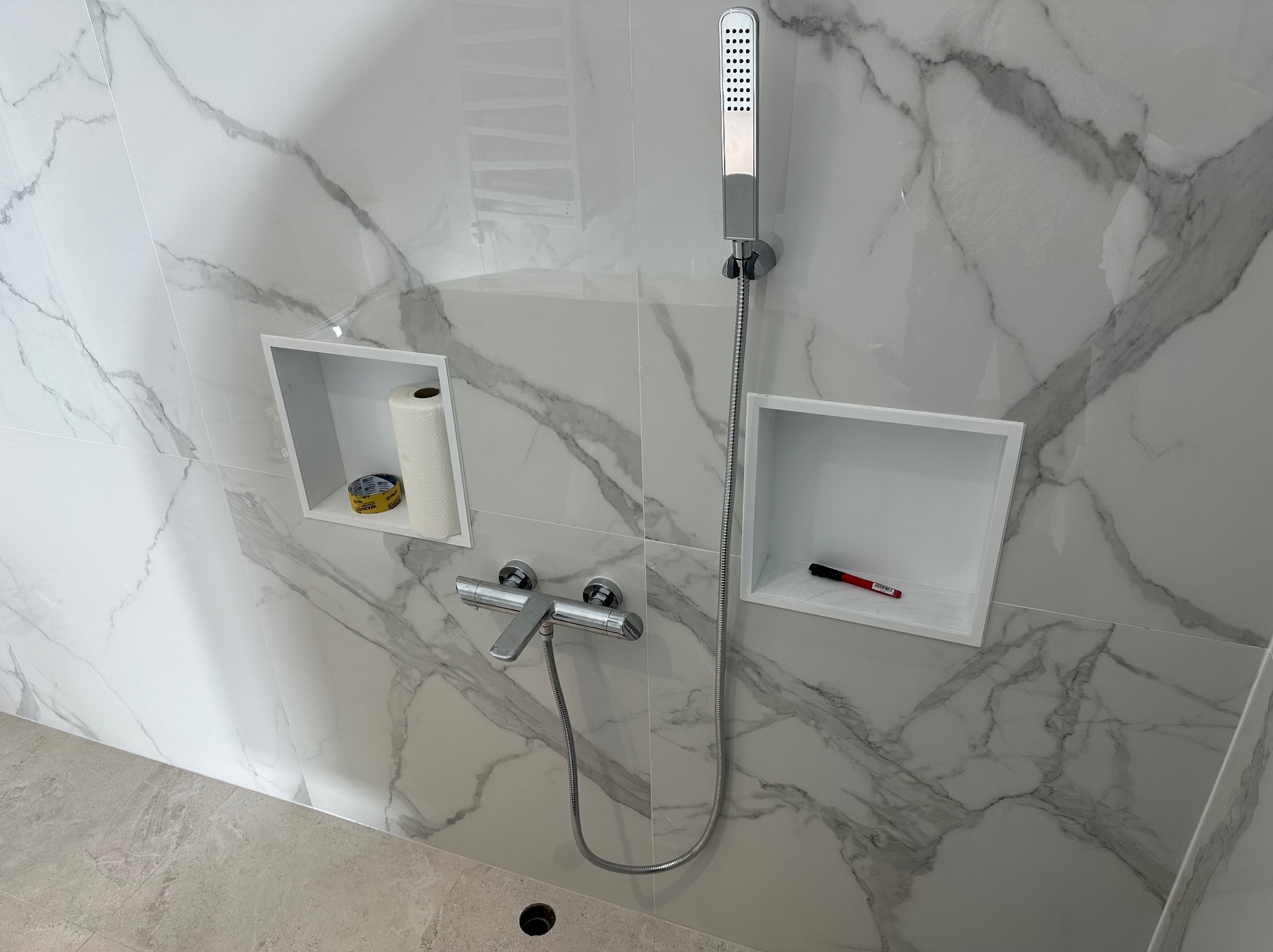 Wykończona łazienka z kabiną prysznicową, baterią i półkami w ścianie wyłożonej białymi płytkami imitującymi marmur. Na półkach akcesoria.