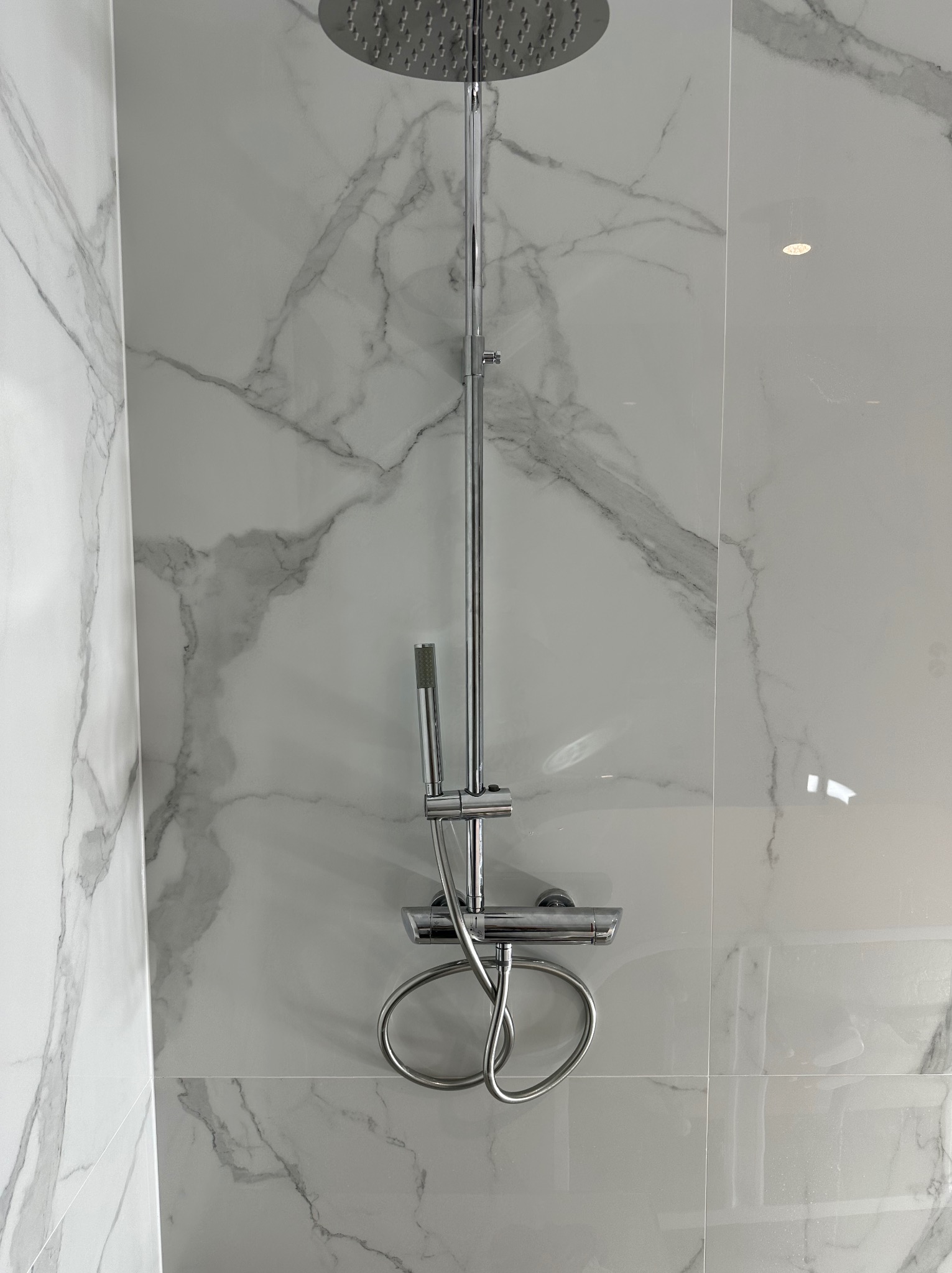 Bateria prysznicowa z deszczownicą na tle białych płytek imitujących marmur. Widoczne chromowane elementy i precyzyjne ułożenie płytek.
