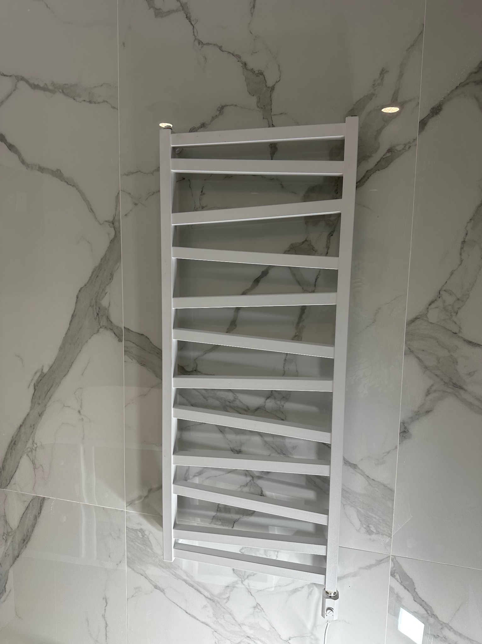 Biały, elektryczny grzejnik łazienkowy drabinkowy na tle marmurowych płytek. Nowoczesny design, ukończony montaż.
