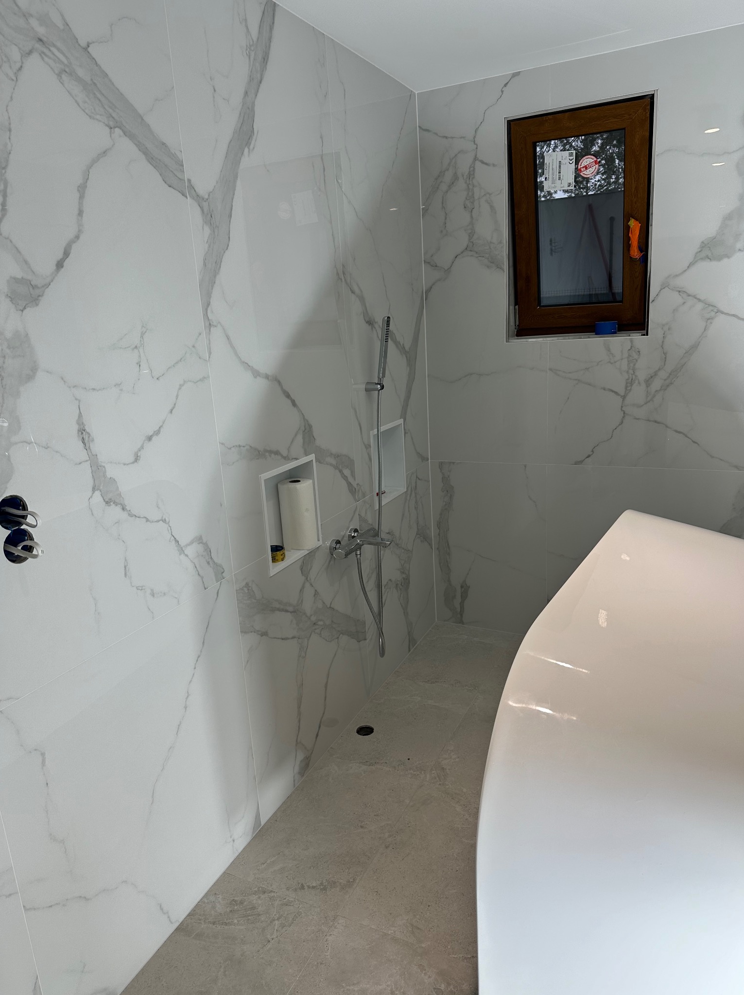 Wykończona łazienka z marmurowymi płytkami, brązowym oknem i białą wanną. Widoczny prysznic i wnęka na papier toaletowy. Nowoczesny design.