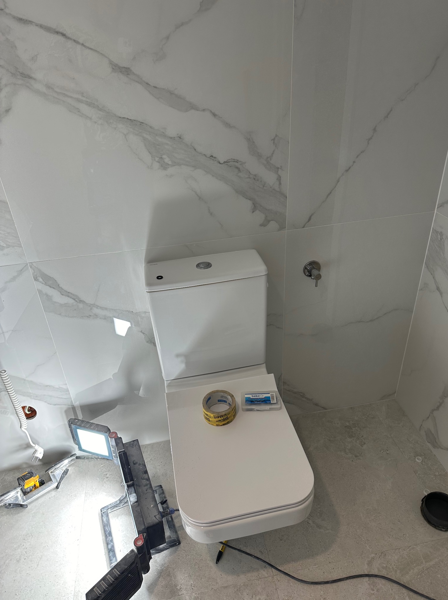 Biała toaleta w łazience z marmurowymi płytkami, oświetlona lampą budowlaną. Widoczna taśma klejąca i opakowanie kołków. Etap wykończenia.
