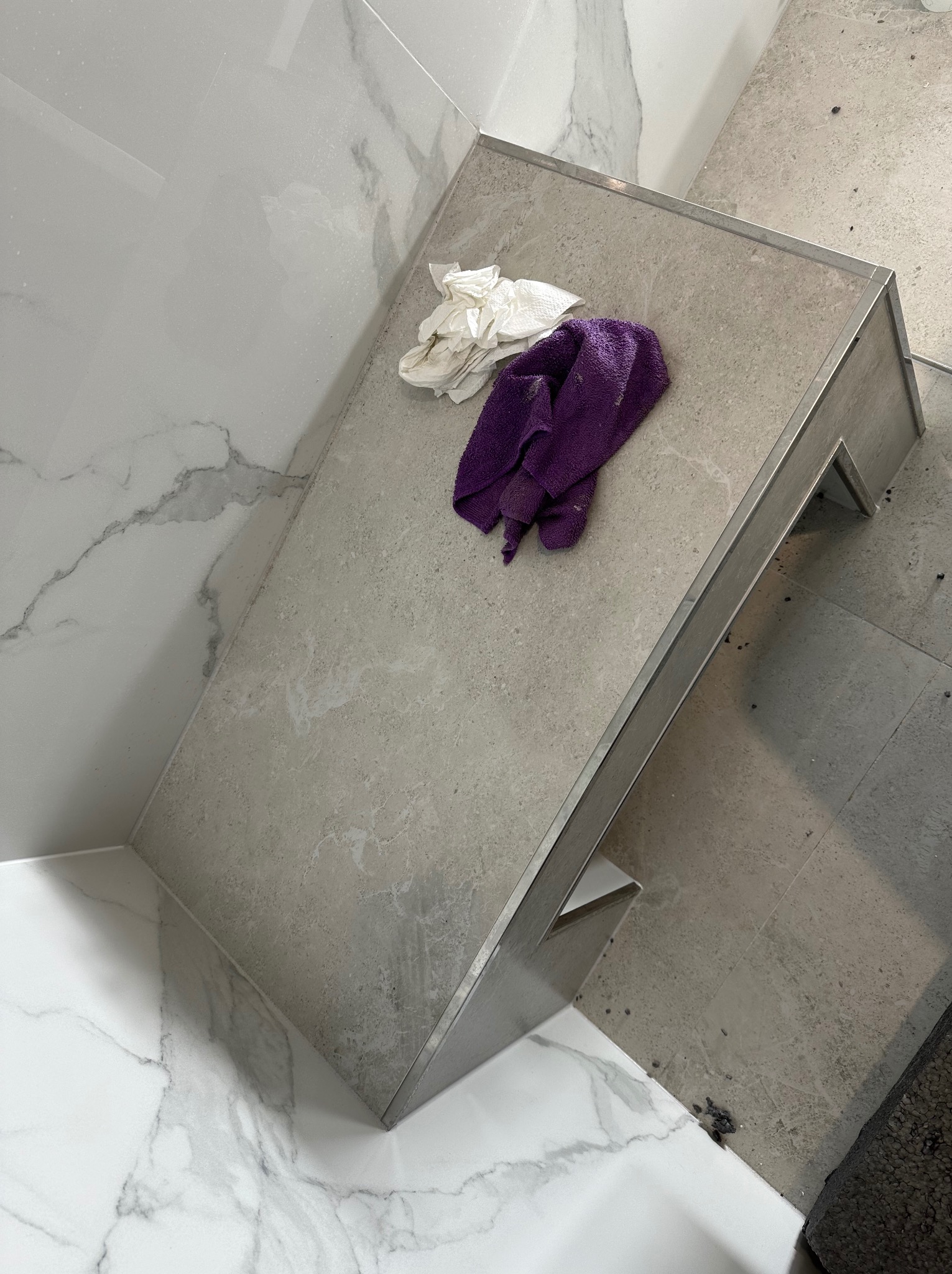 Wykończona półka w łazience z szarych płytek, z widocznymi detalami wykończenia i fugowania. Na półce leżą ściereczki do czyszczenia.