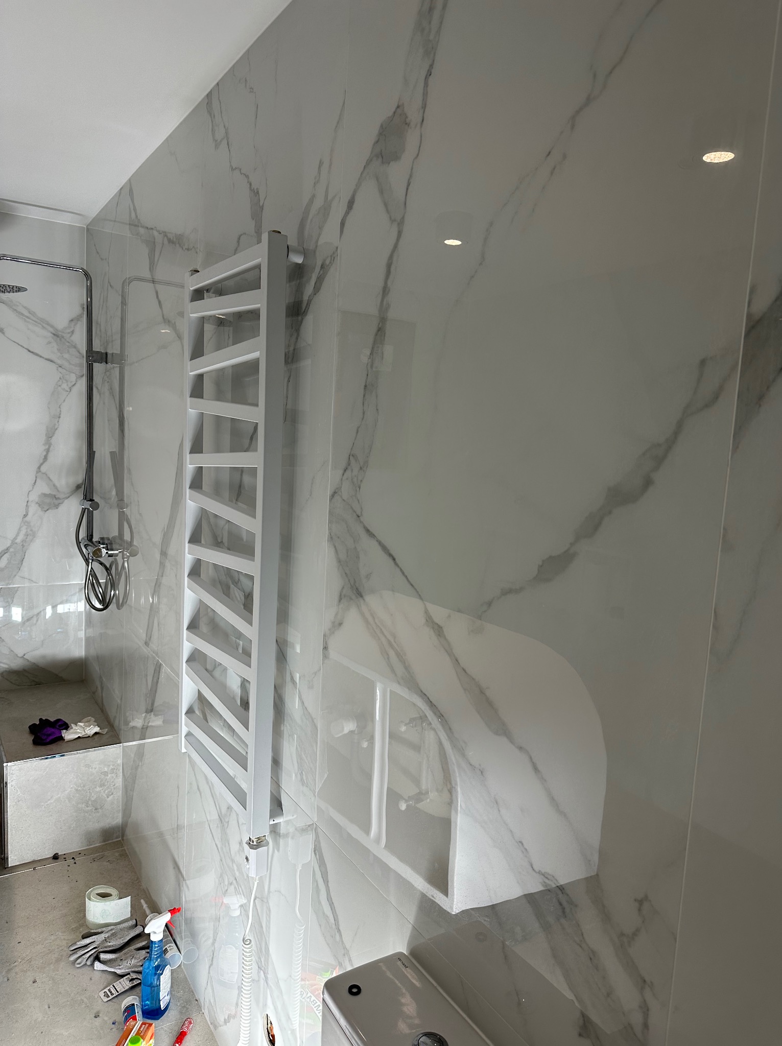 Biała łazienka z marmurowymi płytkami, grzejnikiem drabinkowym i chromowaną armaturą prysznicową. Widoczne narzędzia i akcesoria glazurnicze.