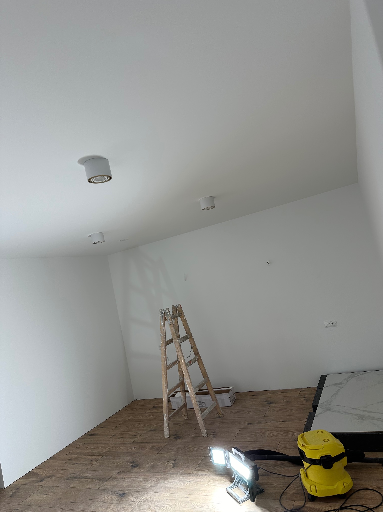 Wnętrze po malowaniu ścian na biało, widoczna drewniana podłoga, drabina malarska, lampa halogenowa i żółty odkurzacz. Oświetlenie sufitowe.