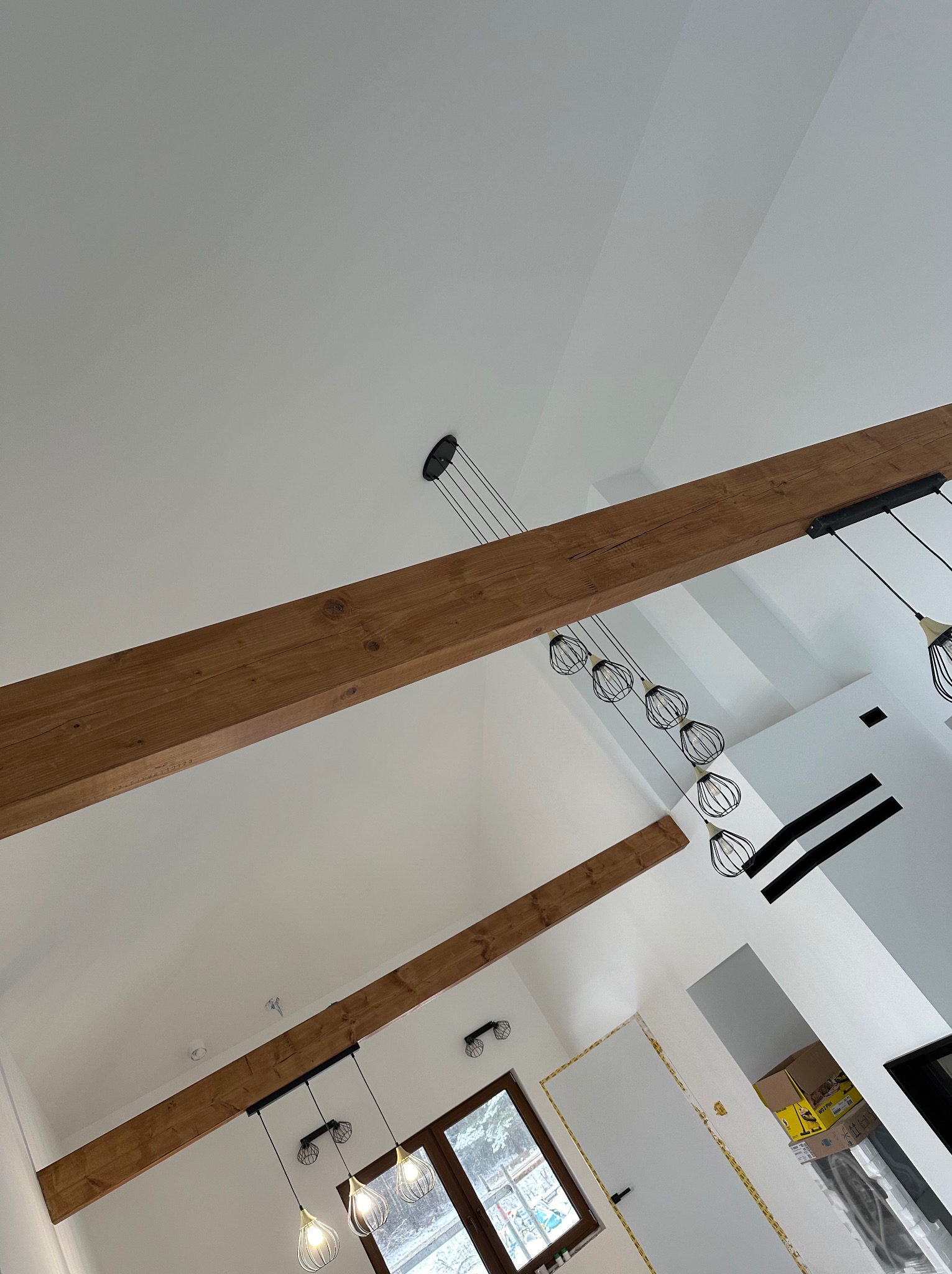 Białe wnętrze ze skośnym sufitem, drewnianymi belkami i nowoczesnym oświetleniem. Widok z dołu ukazuje detale wykończenia i minimalistyczny design.